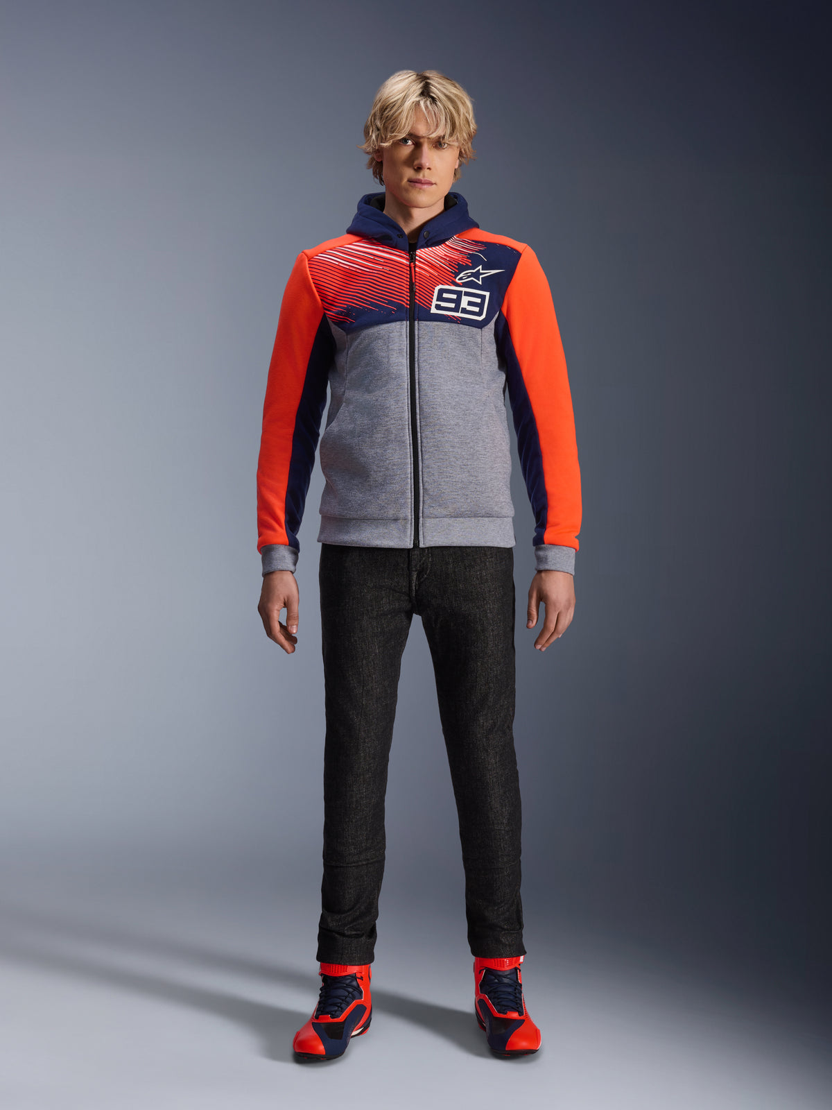 Alpinestars MM93 Chrome V2 Sport Hoodie, Motorrad-Kapuzenjacke, Blau Navy, Grau Melange und Rot Fluo, getragen mit dunkler Motorradhose und Sneakers mit roten Akzenten, mit durchgehendem Reißverschluss und MM93-Logo, lässiges, urbanes, vom Motorsport inspiriertes Design