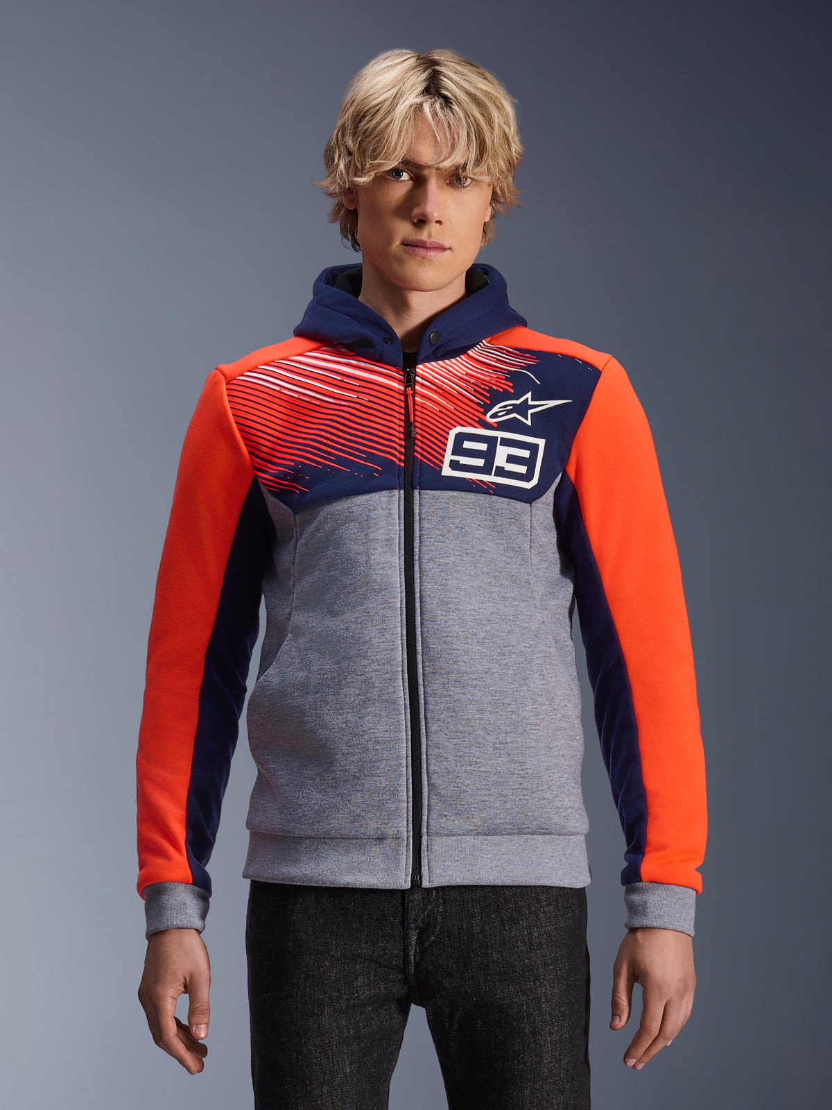 Alpinestars MM93 Chrome V2 Sport Hoodie, Motorrad-Kapuzenjacke mit Reißverschluss, Grau meliert mit dunkelblauem Brustbereich und Kapuze, leuchtend orangefarbene Ärmel, Nummer 93 Design auf der Brust mit Streifenmuster und Stern-Logo, getragen von einem Model mit dunkler Jeans, Marc Márquez Kollektion.