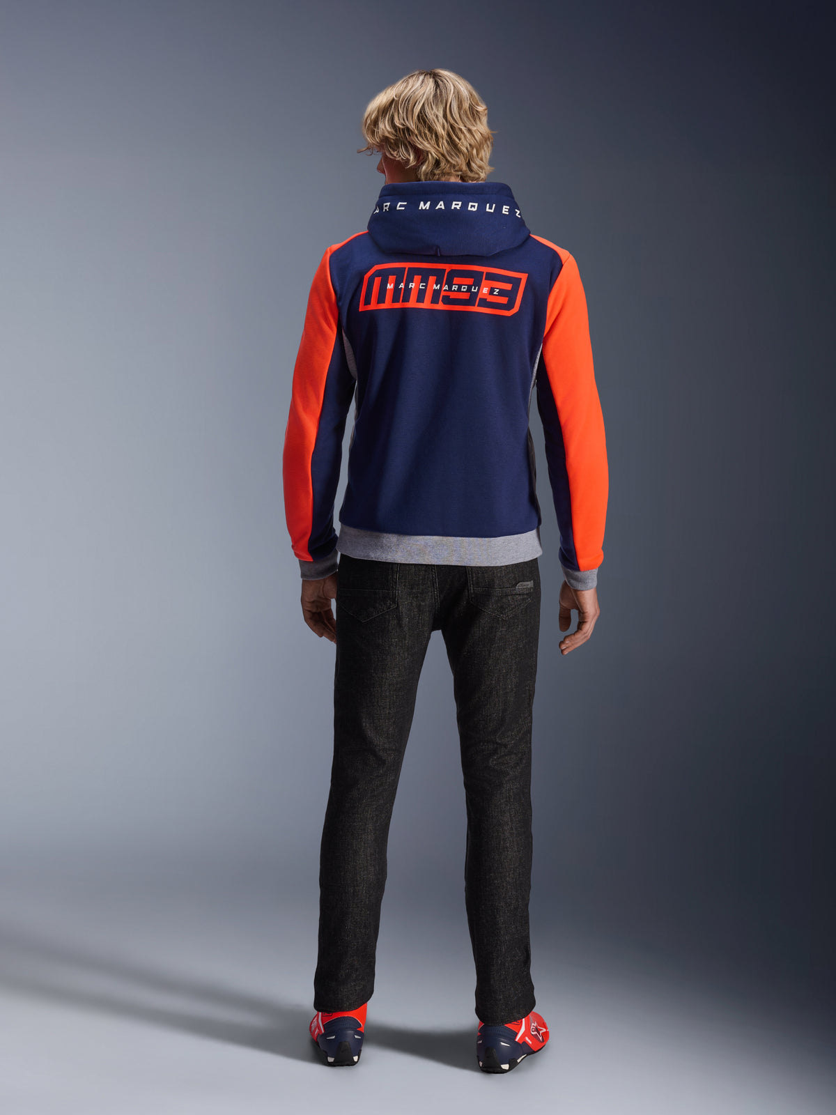 Modell von hinten im Alpinestars MM93 Chrome V2 Sport Hoodie, Dunkelblau mit leuchtend roten Fluo-Ärmeln und Gray-Mel-Details, große MM93- und Marc Marquez-Logos auf dem Rücken und der Kapuze, lässiger Motorrad-Hoodie kombiniert mit einer schwarzen Hose