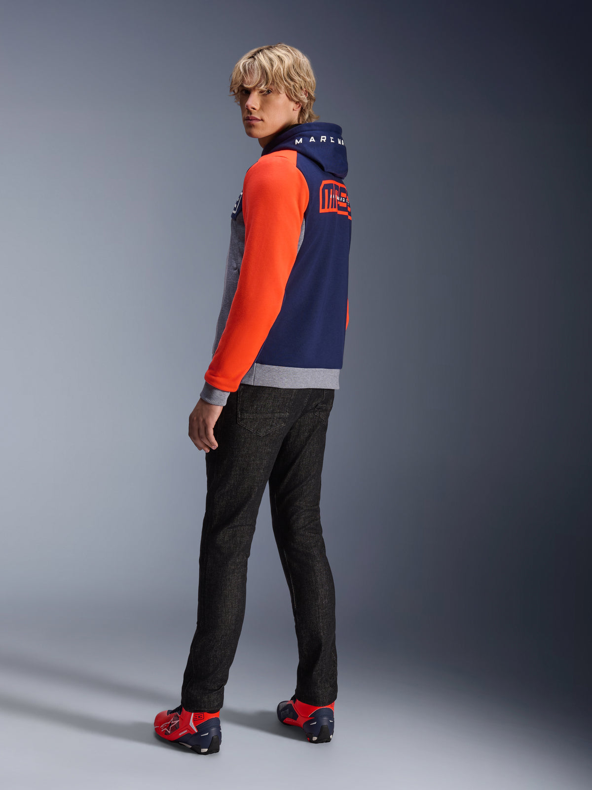 Alpinestars MM93 Chrome V2 Sport Hoodie, Blau Navy Grau Meliert Rot Fluo, Modell trägt Hoodie mit leuchtend roten Ärmeln und MM93 Marc Marquez Branding auf dem Rücken, kombiniert mit dunkler Jeans und roten Motorradschuhen, lässige urbane Motorradausrüstung