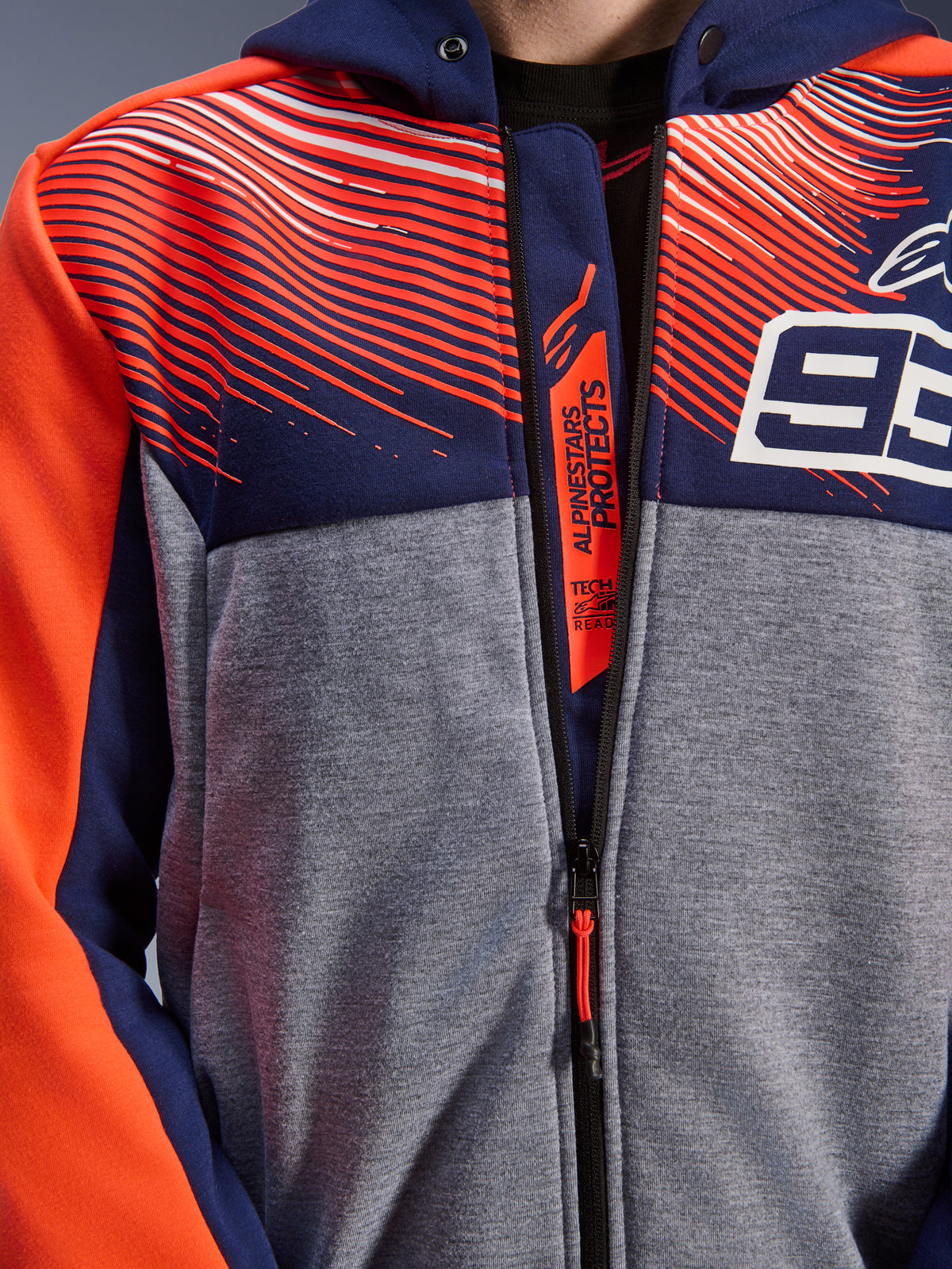 Alpinestars MM93 Chrome V2 Sport Hoodie, Nahaufnahme von Brust und Kragen, Tech-Fleece in Navyblau und Graumeliert mit fluo-roten Grafiken, mit weißem 93-Logo und Alpinestars Protects-Branding auf der inneren Reißverschlussleiste, Tech-Air Ready Motorradbekleidung.