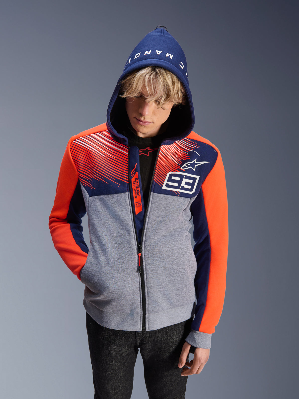 Alpinestars MM93 Chrome V2 Sport Hoodie, Motorrad-Kapuzenjacke, grau melierter Hauptteil mit dunkelblauem Oberbereich und fluoreszierend orangefarbenen Ärmeln, weiße Nummer 93 und Alpinestars Stern-Logo, Marc Marquez Schriftzug auf der Kapuze, getragen von einem männlichen Model mit aufgesetzter Kapuze, urbanes vom Motorsport inspiriertes Design