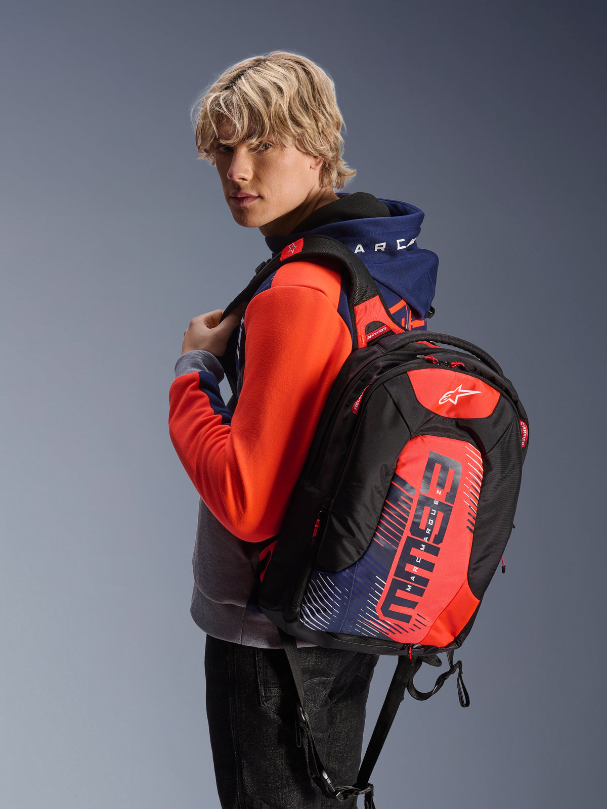 Männliches Model trägt den Alpinestars MM93 Chrome V2 Sport Hoodie in Blue Navy Gray Mel und Red Fluo, kombiniert mit einem passenden MM93-Markenrucksack, lässige urbane Motorradbekleidung mit einer modernen athletischen Silhouette.