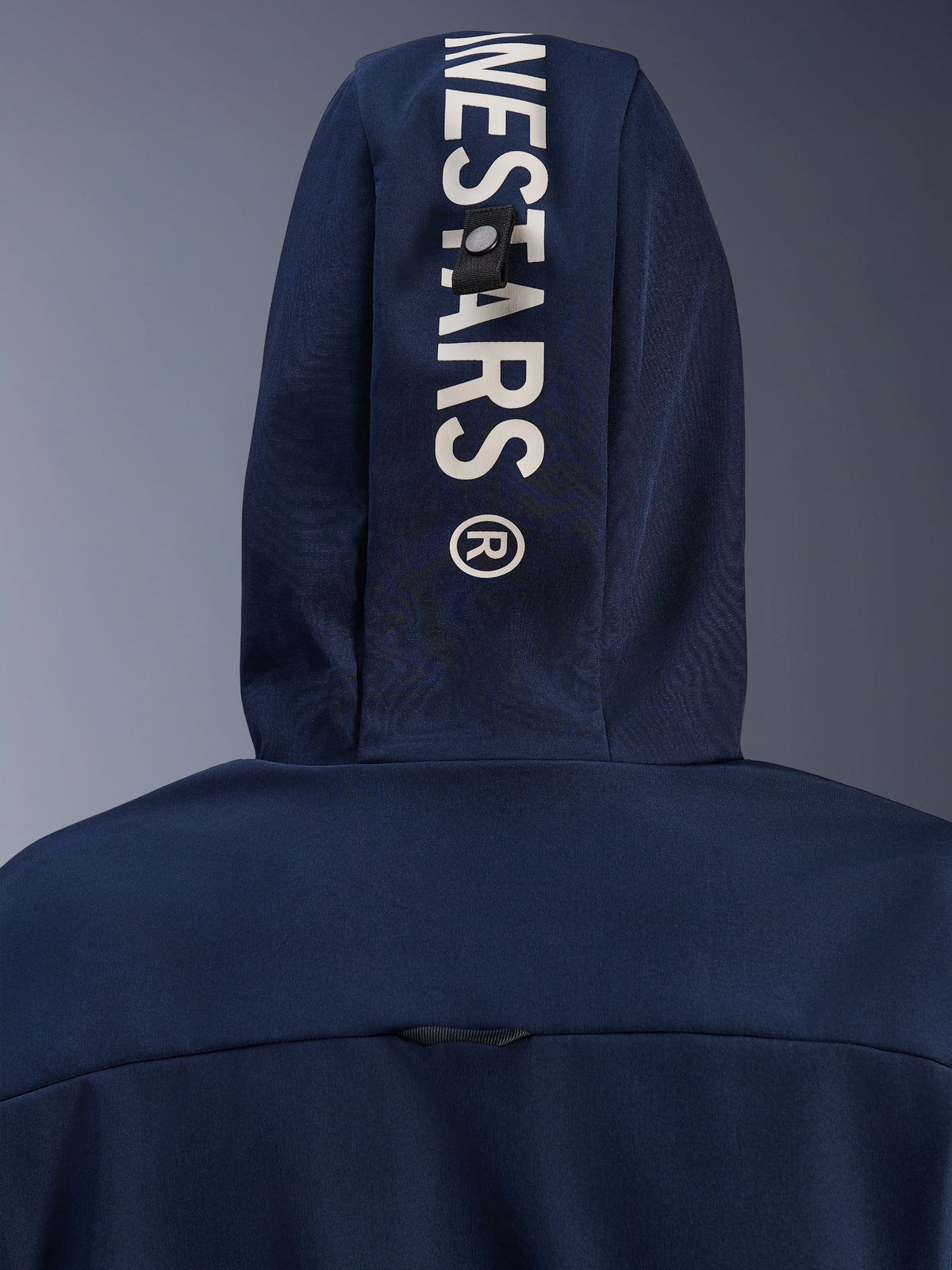 Alpinestars Radium Tech Hoodie in Night Navy Royal Blue, Nahaufnahme der Kapuzenrückseite mit vertikalem weißem Alpinestars-Branding und einem sicheren Befestigungsschlaufen-System, urbane technische Motorradbekleidung.