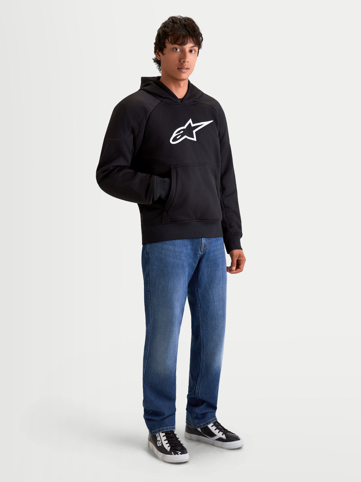 Alpinestars Ageless Kapuzenpullover, urbaner Motorrad-Kapuzenpullover für Herren in Schwarz, Vorderansicht an Model in lässigen Jeans, mit weißem Astar-Logo auf der Brust und internen Abrisssicherungen