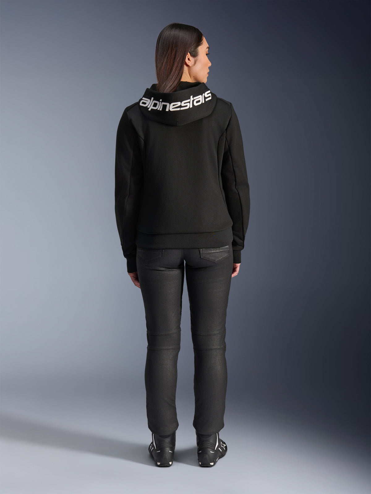 Rückansicht eines Models im Alpinestars Stella Chrome V2 Sport Hoodie in Schwarz, mit einem markanten weißen Alpinestars-Logo auf der Kapuze, kombiniert mit einer schwarzen Motorradhose und Motorradstiefeln, urbane Sport-Motorradbekleidung für Damen