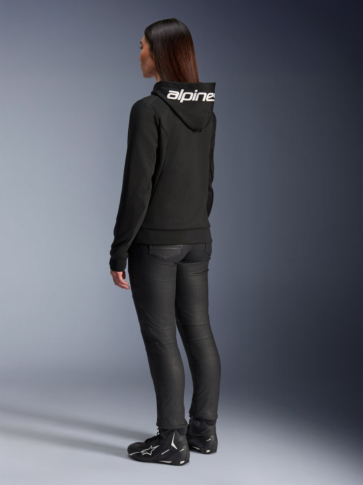 Frau trägt Alpinestars Stella Chrome V2 Sport Hoodie, Rückansicht, schwarzer Motorrad-Hoodie mit weißem Alpinestars-Logo auf der Kapuze, kombiniert mit schwarzer technischer Fahrhose und schwarzen Motorradschuhen, lässige urbane Pendlerausrüstung
