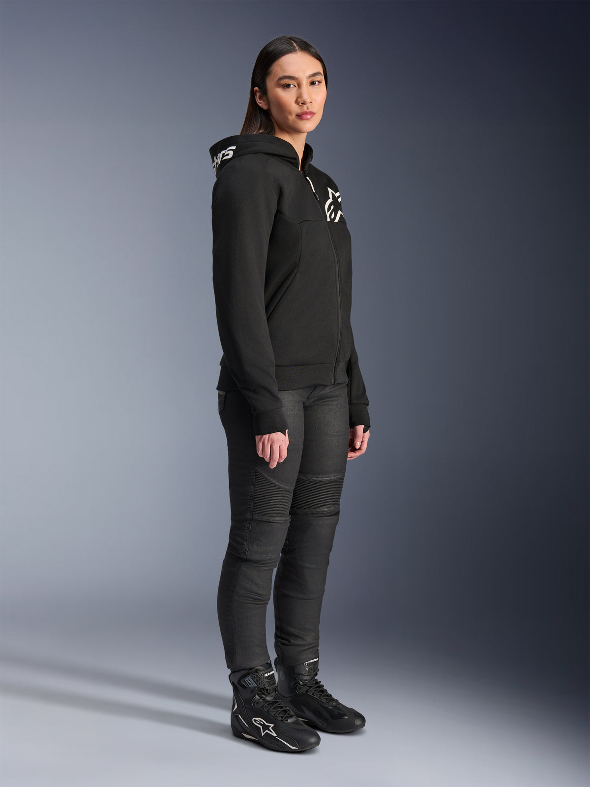 Frau im Alpinestars Stella Chrome V2 Sport Hoodie in Schwarz mit weißem Alpinestars-Branding, kombiniert mit schwarzer Motorradhose und Stiefeln, urbane Motorradausrüstung für Damen mit lässiger, sportlicher Passform
