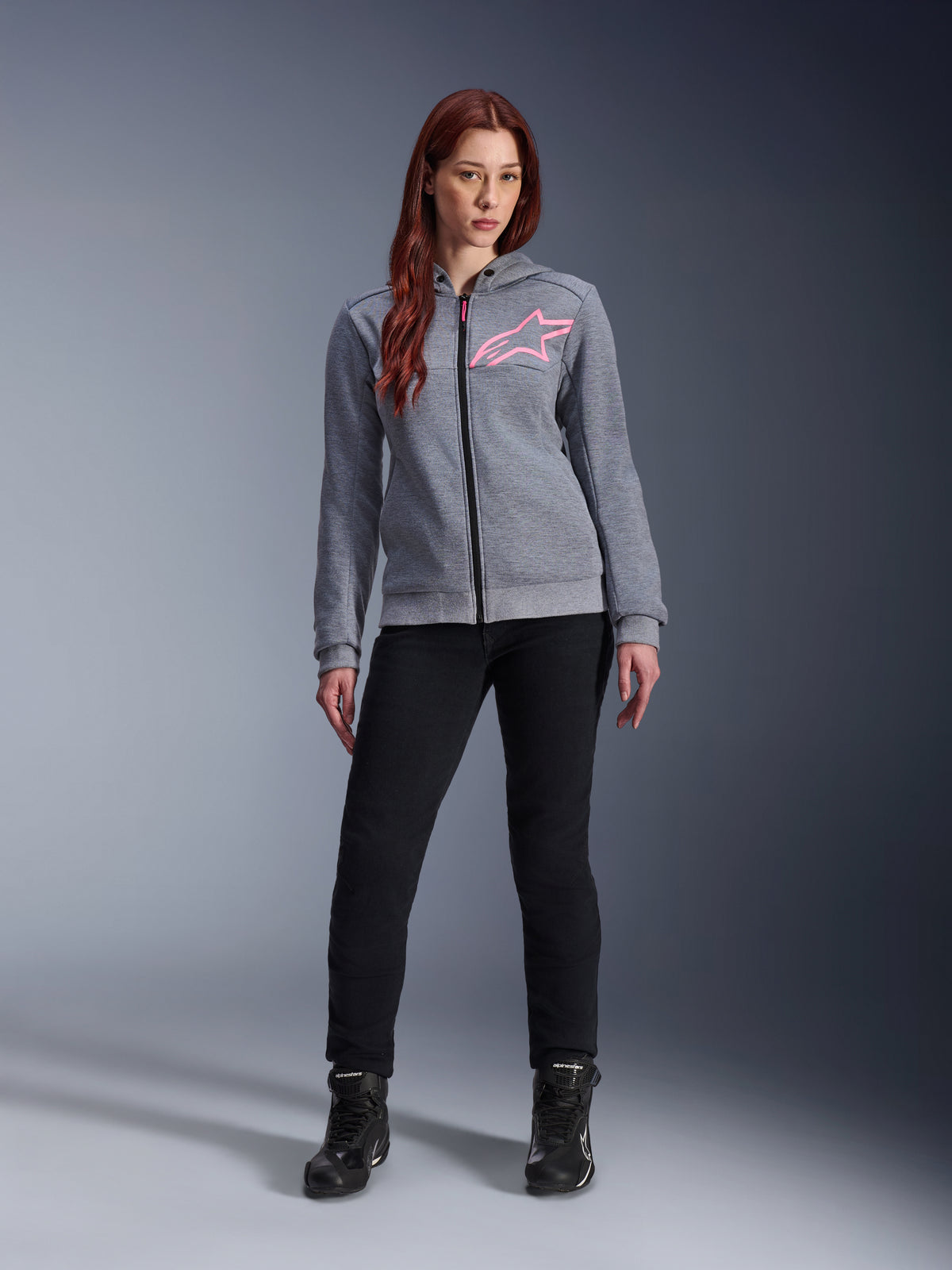 Alpinestars Stella Chrome V2 Sport Hoodie, Gray Heather Pink Fluo, getragen von einer Frau mit schwarzer Hose und Stiefeln, mit einem pinkfarbenen Alpinestars Stern-Logo auf der Brust, Urban-Motorradbekleidung mit durchgehendem Reißverschluss, leichtes Tech-Fleece mit lässiger, sportlicher Passform.