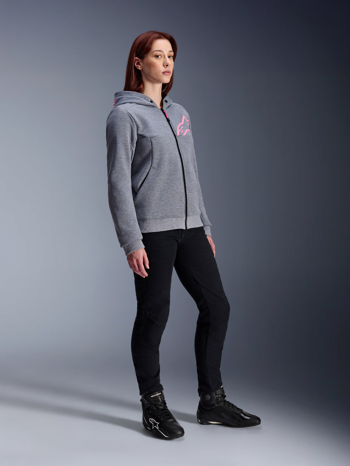 Alpinestars Stella Chrome V2 Sport Hoodie, Motorrad-Kapuzenjacke für Damen, Grau-Meliert und Neon-Pink, getragen mit schwarzer Motorradhose und schwarzen Stiefeln, lässige urbane Motorradausrüstung