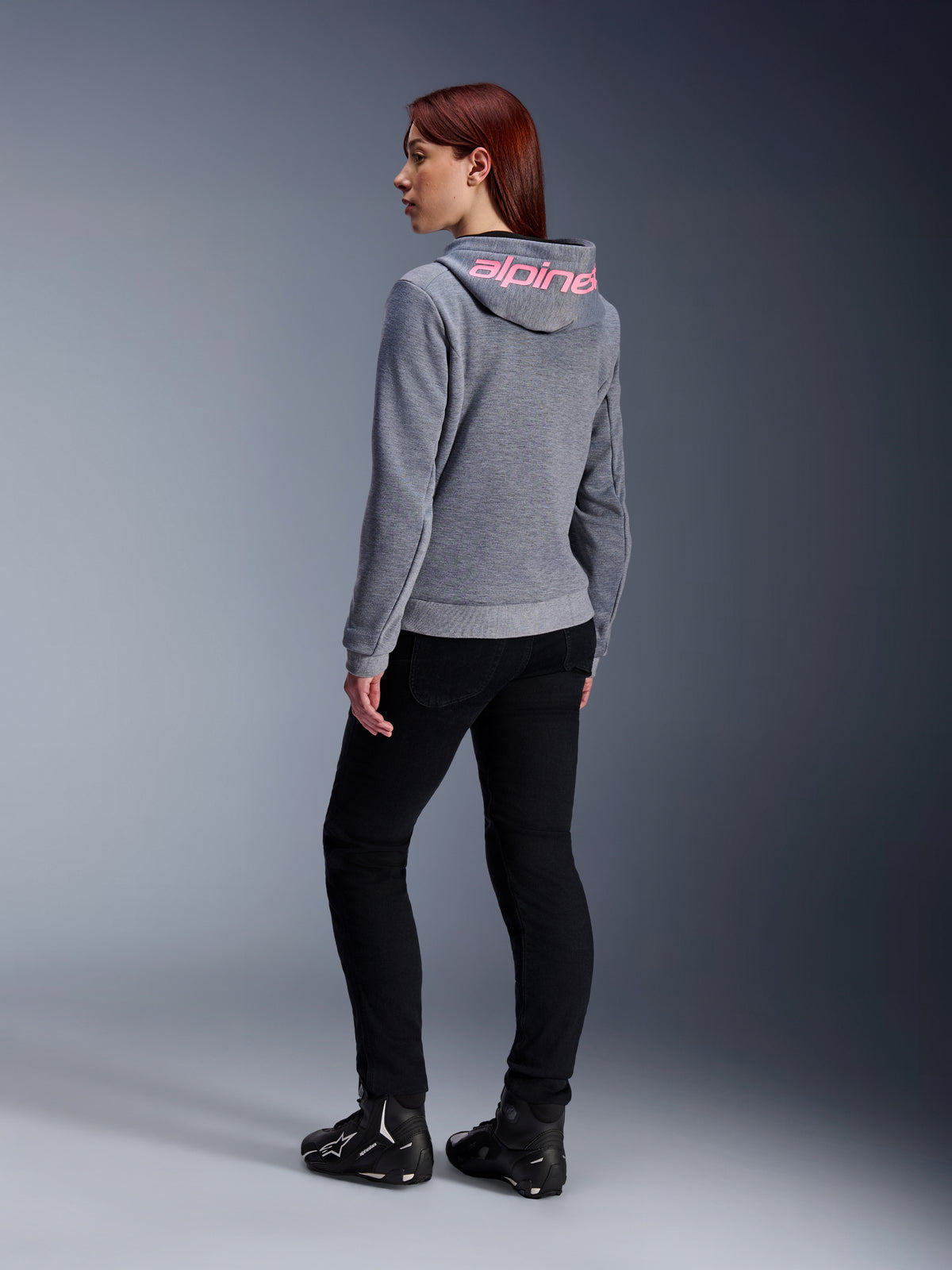 Alpinestars Stella Chrome V2 Sport Hoodie, Damen-Motorrad-Hoodie, Grau meliert mit pinkem Fluo-Branding auf der Kapuze, Rückansicht, kombiniert mit schwarzer Hose und Motorradschuhen, lässiger urbaner Street-Style mit Motorradschutz