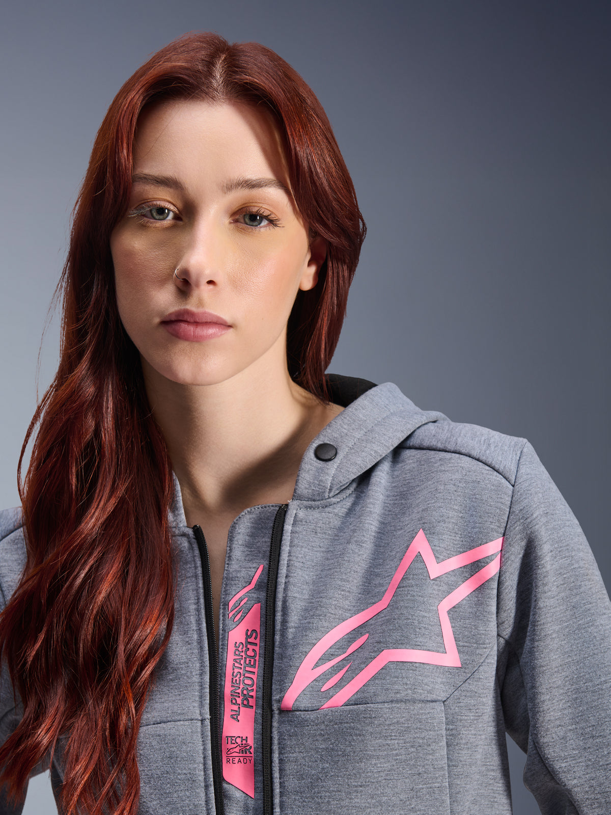 Frau trägt Alpinestars Stella Chrome V2 Sport Hoodie, Grau Meliert Pink Fluo, Nahaufnahme mit dem pinkfarbenen Alpinestars Stern-Logo und Tech-Air Ready Branding, urbane Motorradbekleidung.