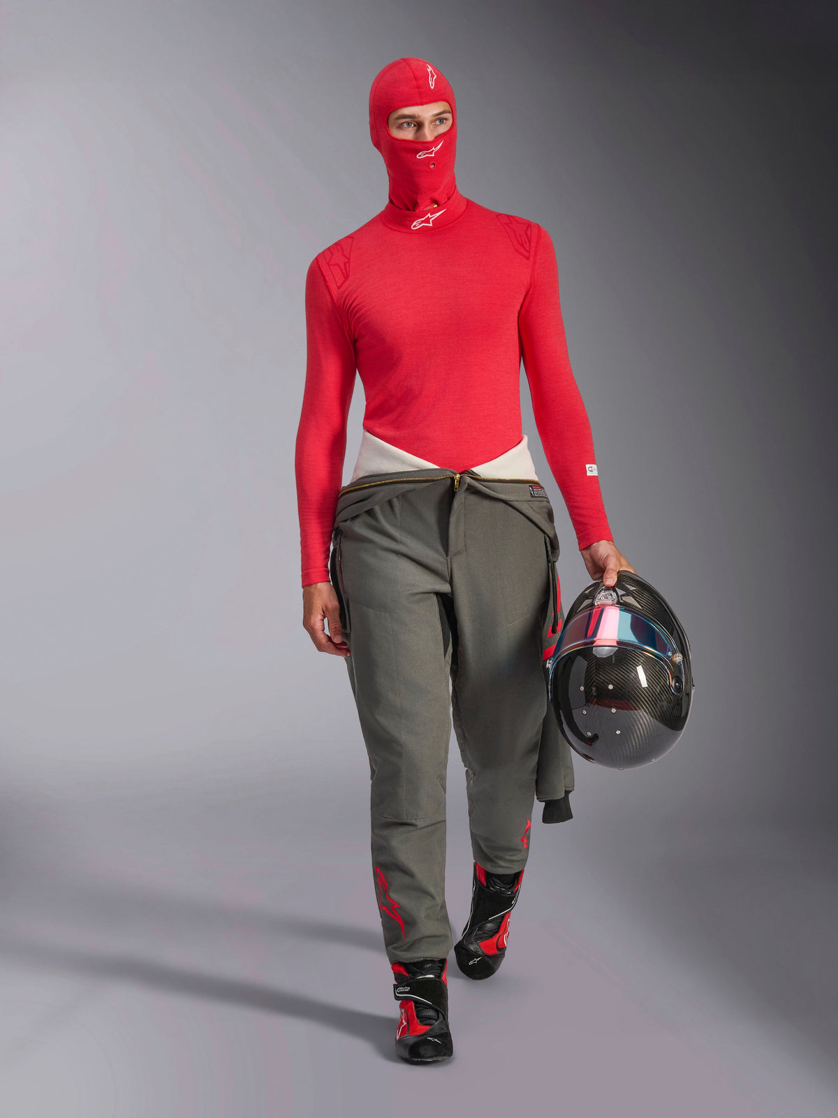 Rennfahrer mit Alpinestars ZX Evo V3 Sturmhaube in Rot, passender roter langärmeliger Base Layer, graue Rennhose, schwarz-rote Rennstiefel, trägt einen Carbon-Helm, professionelle Automobil-Rennausrüstung