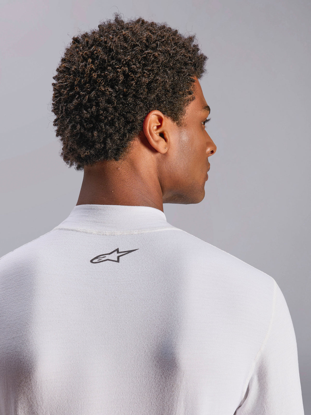 Alpinestars ZX Evo V3 Oberteil, Langarm-Rennsport-Unterwäsche, Weiß, Nahaufnahme des Nackenbereichs mit schwarzem Alpinestars-Logo, technisches flammhemmendes Material, getragen von einem Modell mit dunklem lockigem Haar
