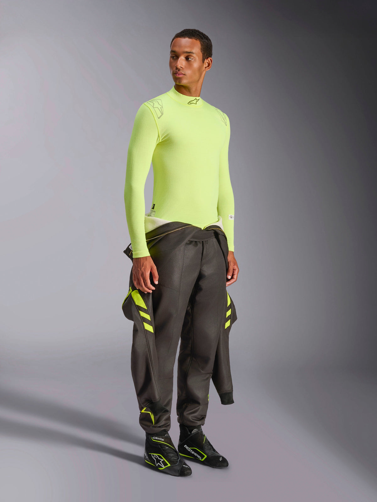Alpinestars ZX Evo V3 Top - Long Sleeve, Yellow Fluo, worn by a model with a dark grey racing suit tied at the waist and black racing boots, flame-resistant motorsport base layer, lightweight jacquard mesh construction, professional auto racing gear