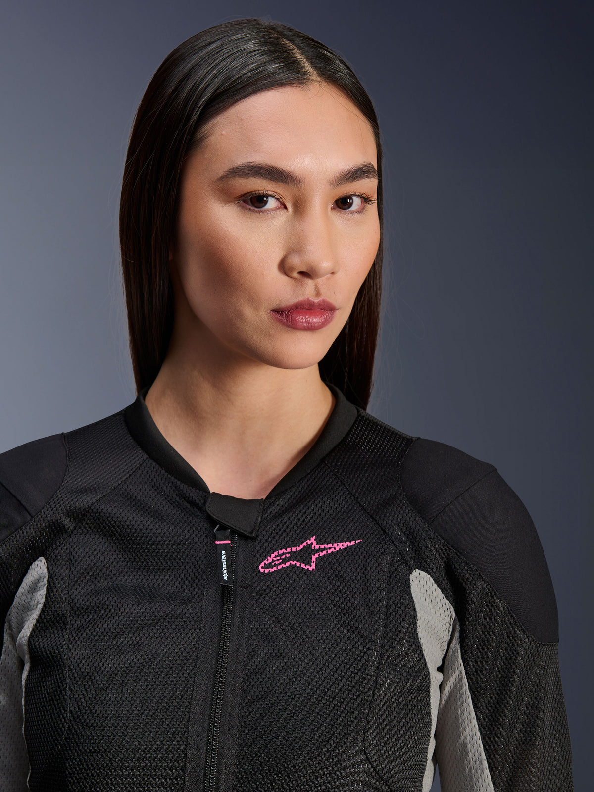 Frau in der Alpinestars Stella Troop-Air Jacke, Schwarz, Aschegrau und Pink Fluo, Nahaufnahme mit Fokus auf die technische Mesh-Konstruktion und das pinkfarbene Brustlogo, stylische urbane Motorradausrüstung