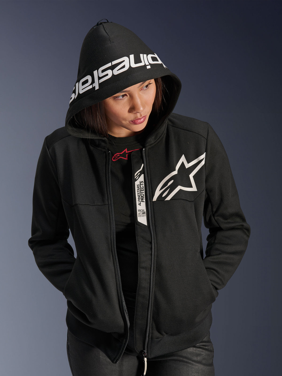 Alpinestars Stella Chrome V2 Sport Hoodie für Damen, Schwarz und Weiß, Motorrad-Kapuzenjacke mit weißem Stern-Logo auf der Brust und Alpinestars-Branding am Kapuzenrand, Frau mit aufgesetzter Kapuze, lässiger urbaner Sport-Style mit Tech-Air Ready-Details
