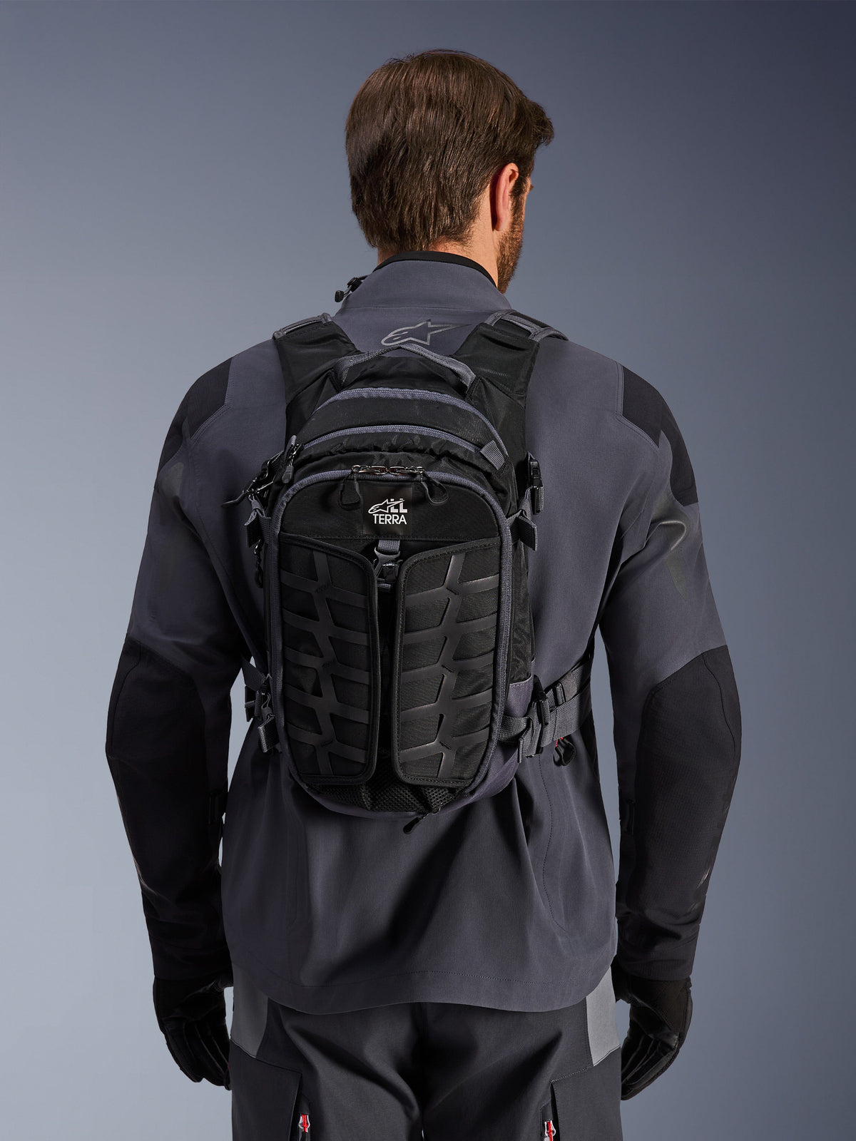 Alpinestars AT-15 Rucksack, Adventure-Motorradrucksack, Black Deep Gray, getragen über einer grauen technischen Motorradjacke, Rückansicht mit „ALL TERRA“-Branding, ergonomisches Design mit verstärkten, strukturierten Paneelen und sicheren Gurten.