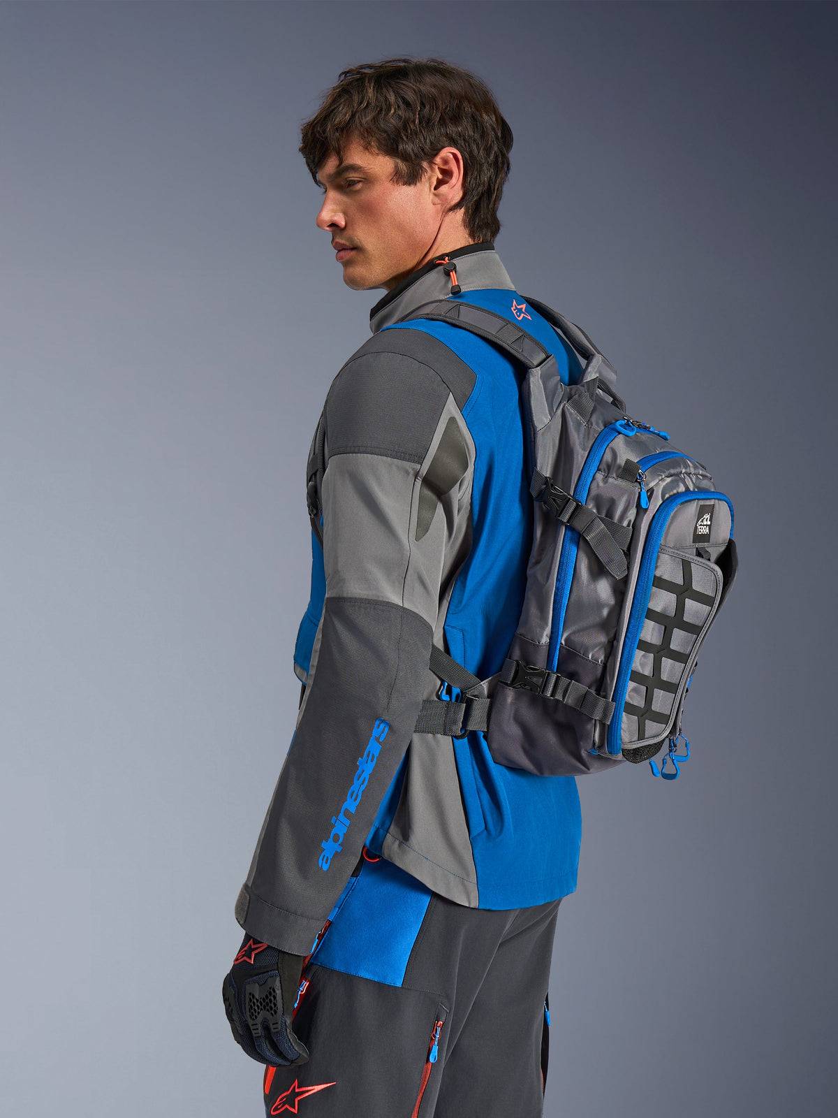 Alpinestars AT-15 Rucksack, Motorrad-Adventure-Rucksack, Dunkelgrau mit blauen Akzenten, getragen von einem Fahrer in farblich abgestimmter grau-blauer technischer Fahrbekleidung, mit MOLLE-Panel und robuster Konstruktion für Offroad-Abenteuer.