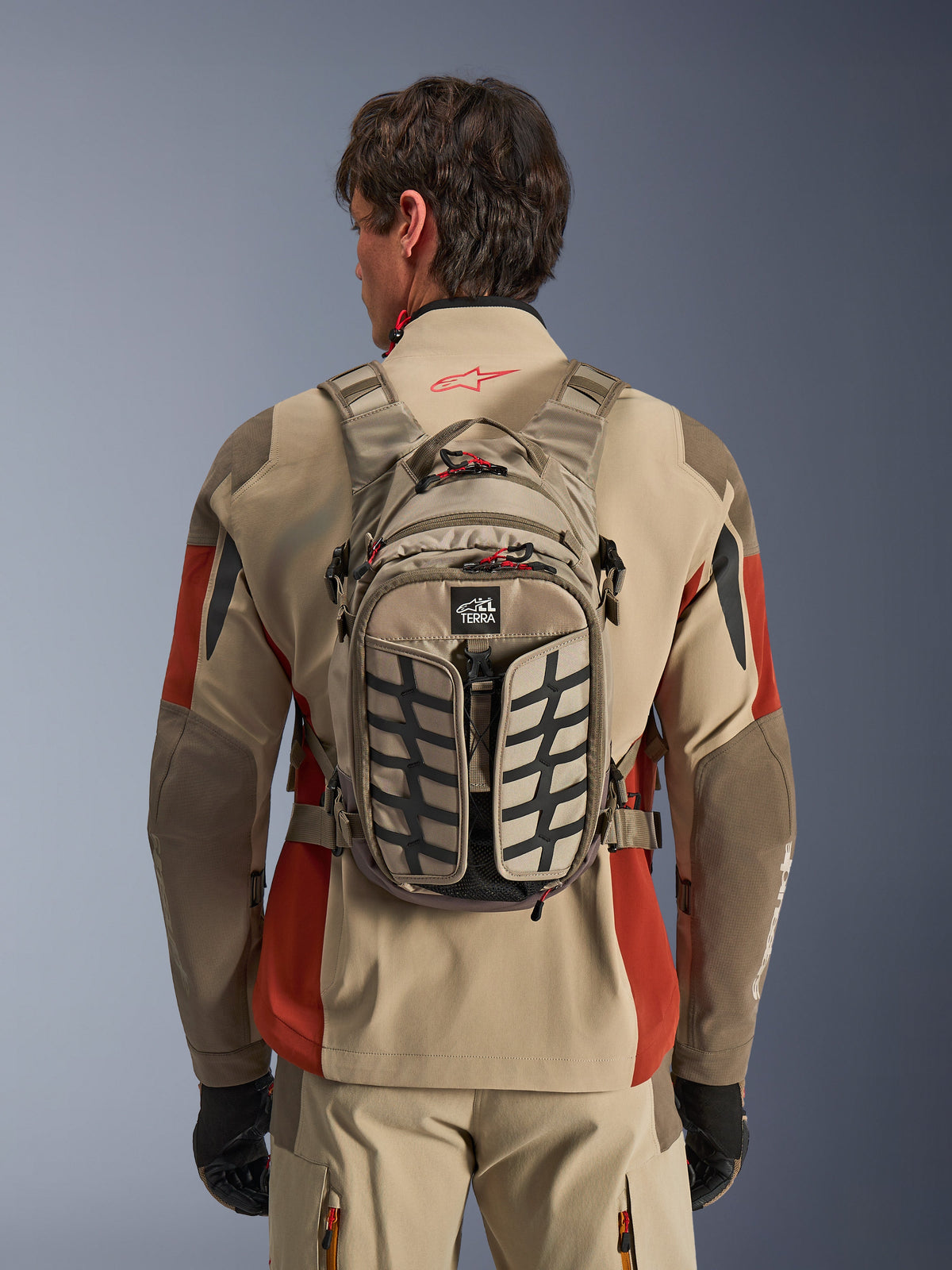 Alpinestars AT-15 Back Pack, Aluminum Walnut, technischer Adventure-Motorradrucksack, getragen von einem Fahrer in beige-roter Jacke, mit Reifenprofil-Muster, TERRA-Logo, roten Reißverschluss-Laschen und verstellbaren Gurten
