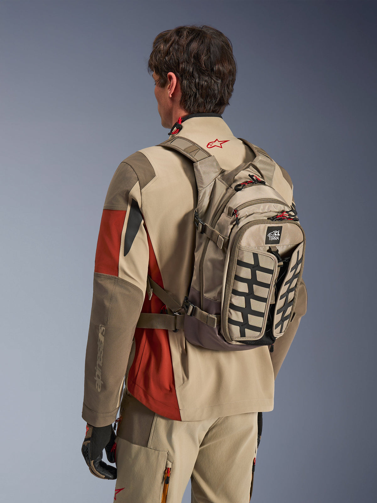 Alpinestars AT-15 Rucksack in Aluminum Walnut, technischer Motorradrucksack, getragen von einem Fahrer in passender Adventure-Ausrüstung, mit einer Textilkonstruktion in Hellbraun und Braun, schwarzen strukturellen Akzenten und roten Reißverschluss-Zippern, konzipiert für Offroad- und Adventure-Fahrten.