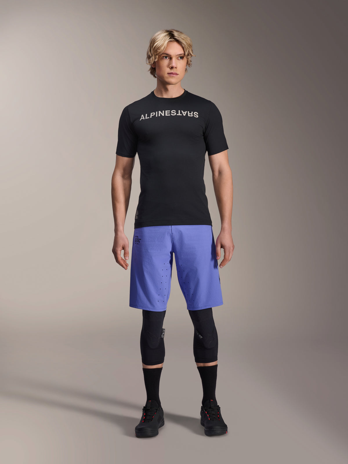 Alpinestars A-Aria Switch Trikot, Kurzarm-MTB-Trikot, Schwarz mit gespiegeltem weißem Alpinestars-Logo, getragen von einem männlichen Model mit blauen Radshorts und schwarzen Knieschonern, atmungsaktive Performance-Bekleidung für Trail-Fahrten