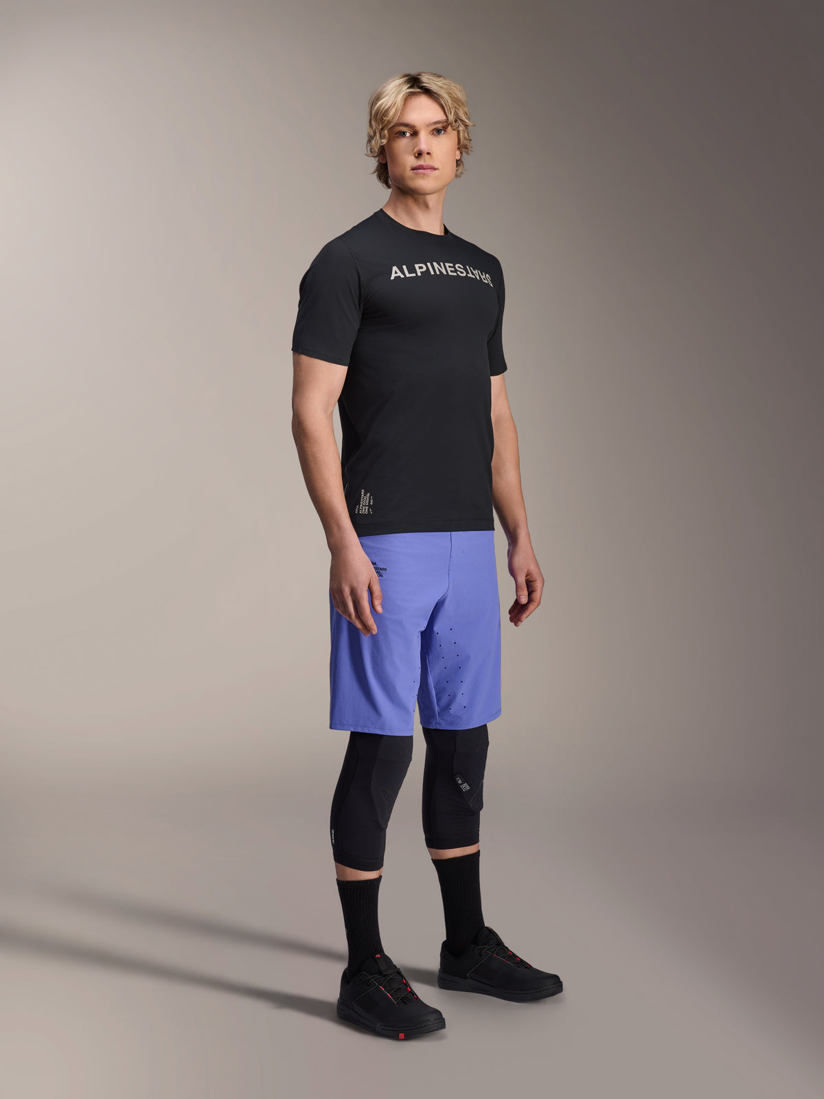 Männliches Model trägt das Alpinestars A-Aria Switch Kurzarmtrikot in Schwarz mit weißem Branding auf der Brust, kombiniert mit hellblauen Mountainbike-Shorts, schwarzen Knieschonern und schwarzen Radschuhen, Ganzkörper-Studioaufnahme für Performance-MTB-Ausrüstung