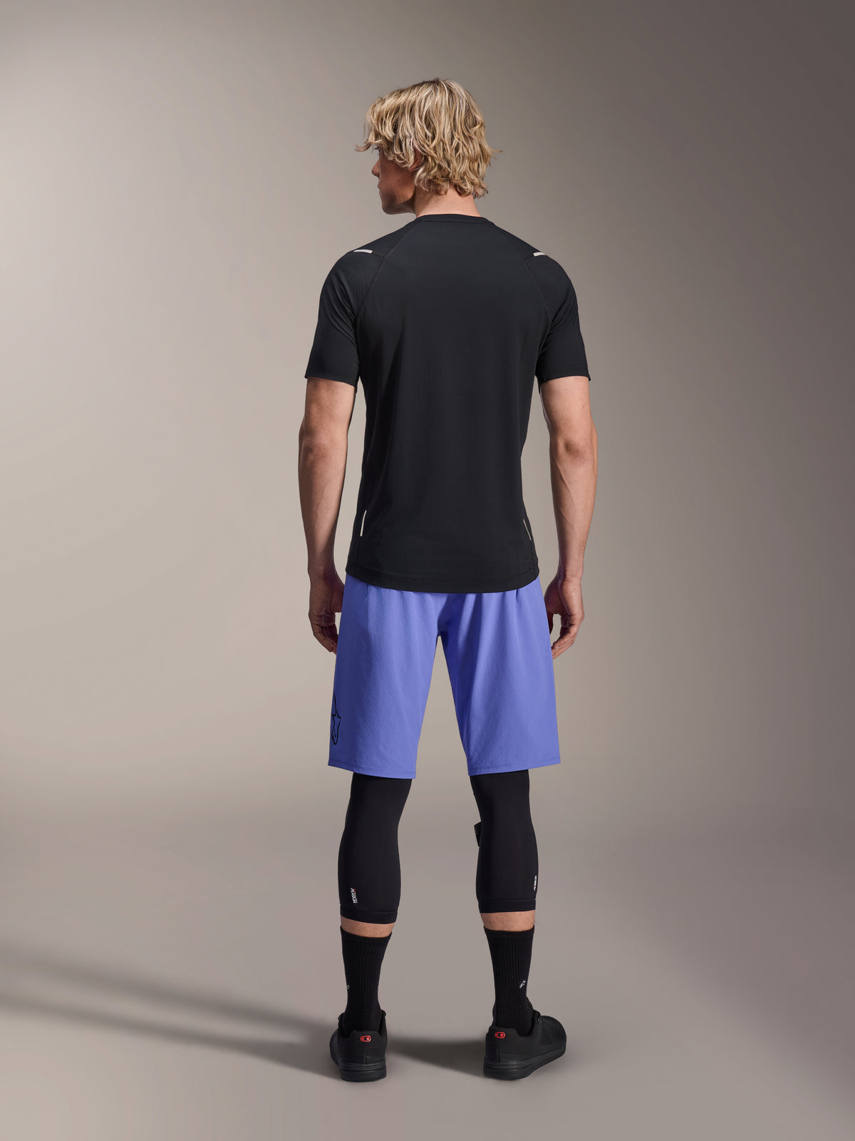 Männlicher Fahrer in Alpinestars A-Aria Lite Shorts, MTB-Shorts, Baja Blue, Rückansicht, kombiniert mit einem schwarzen Kurzarmtrikot, schwarzen Knieprotektoren und schwarzen Radschuhen, Performance-Mountainbike-Bekleidung