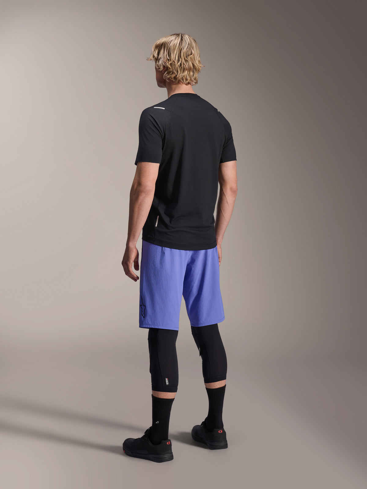 Alpinestars A-Aria Switch Trikot, kurzärmeliges MTB-Trikot, Schwarz, Rückansicht am Modell mit blauen Shorts und schwarzen Knieprotektoren, Performance-Radbekleidung für Trail-Fahrten
