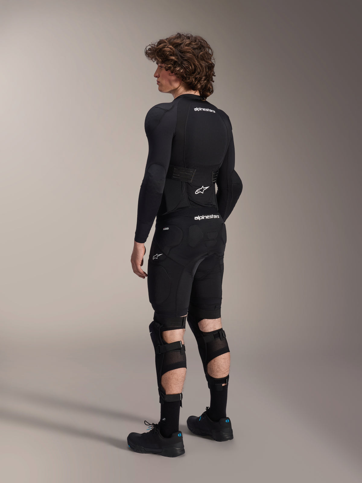 Modell trägt Alpinestars A-Impact Plasma Pro Knie-Schienbein-Protektoren in der Rückansicht, Schwarz und Weiß, zeigt den Dreifach-Riemenverschluss und belüftete Mesh-Einsätze, kombiniert mit einer kompletten schwarzen Mountainbike-Schutzausrüstung inklusive langärmeligem Baselayer und gepolsterten Shorts