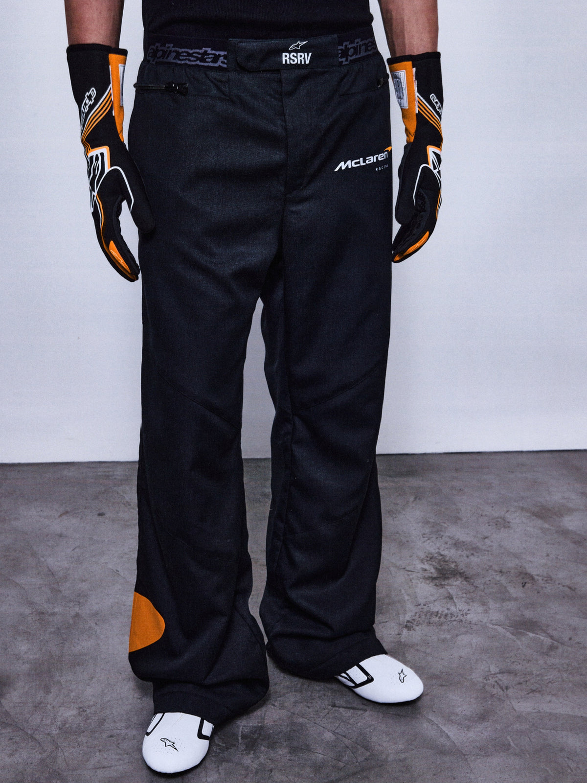 RSRV Arena Suit Pants, Black Racing Pants with McLaren and Alpinestars Logos, Orange Side Patches, Premium Motorsport Apparel, Worn with Black and Orange Gloves and White Racing Shoes