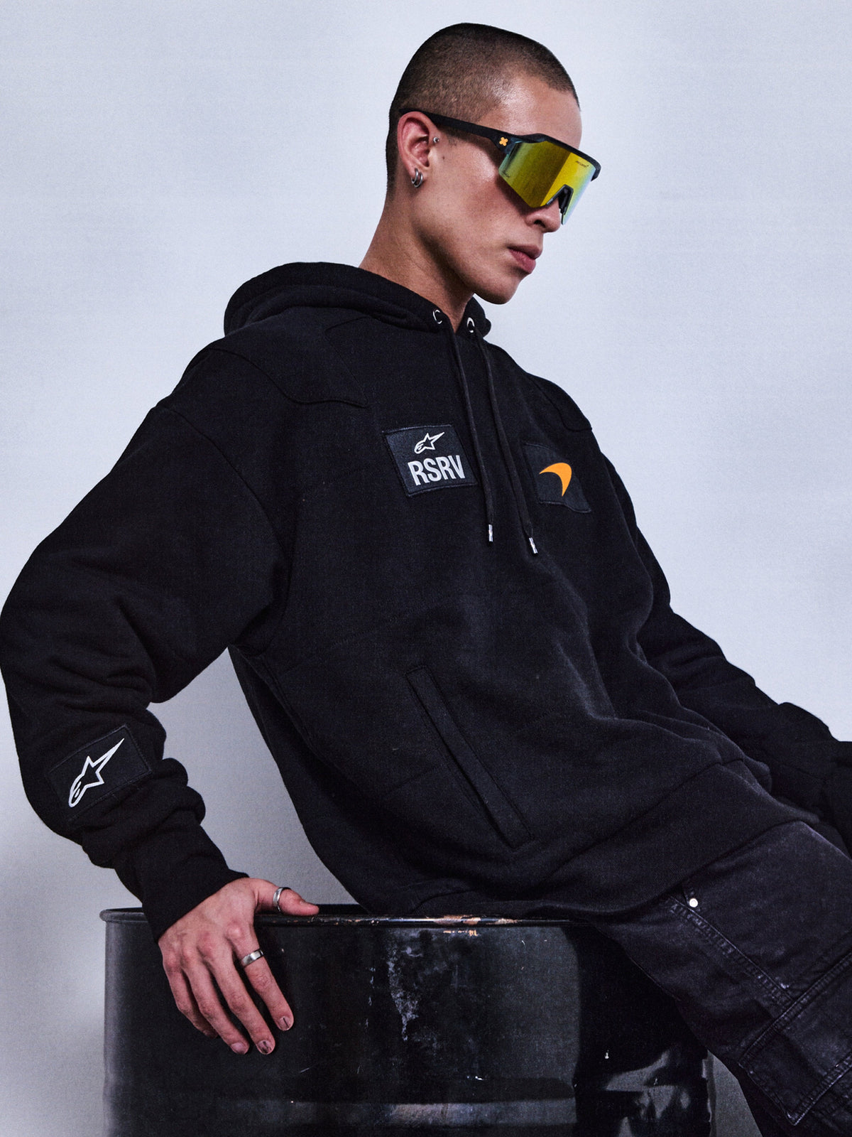 Alpinestars RSRV Rival Hoodie, Schwarzer Premium-Hoodie mit RSRV-Logo und Patches, Unisex-Streetwear, Person mit reflektierender Sonnenbrille auf schwarzem Metallfass, urbaner Lifestyle-Look