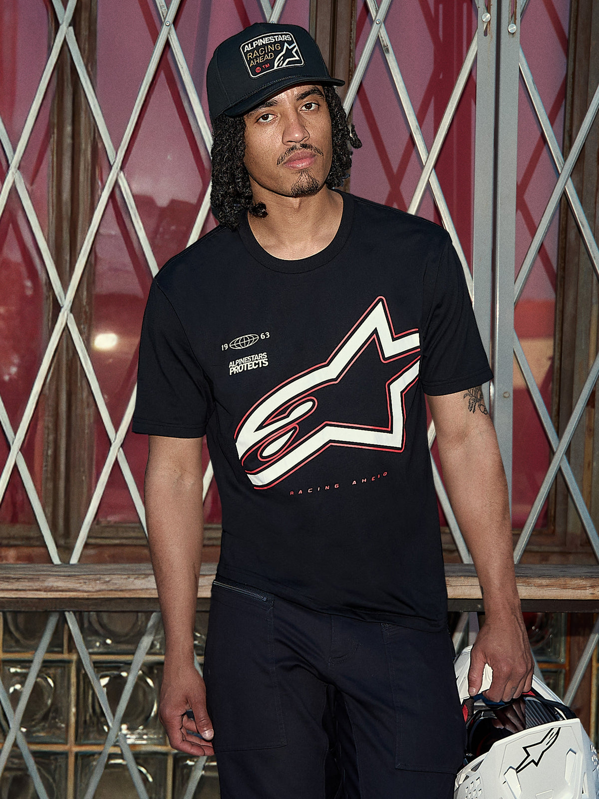 Alpinestars Authority CSF T-Shirt, kurzärmeliges T-Shirt, Schwarz, großer weißer und roter Astars-Logo-Umriss mit „Alpinestars Protects“-Grafik, Lifestyle-Aufnahme eines Mannes mit schwarzer Trucker-Kappe, der einen weißen Helm hält, lässige Motorsport-Streetwear