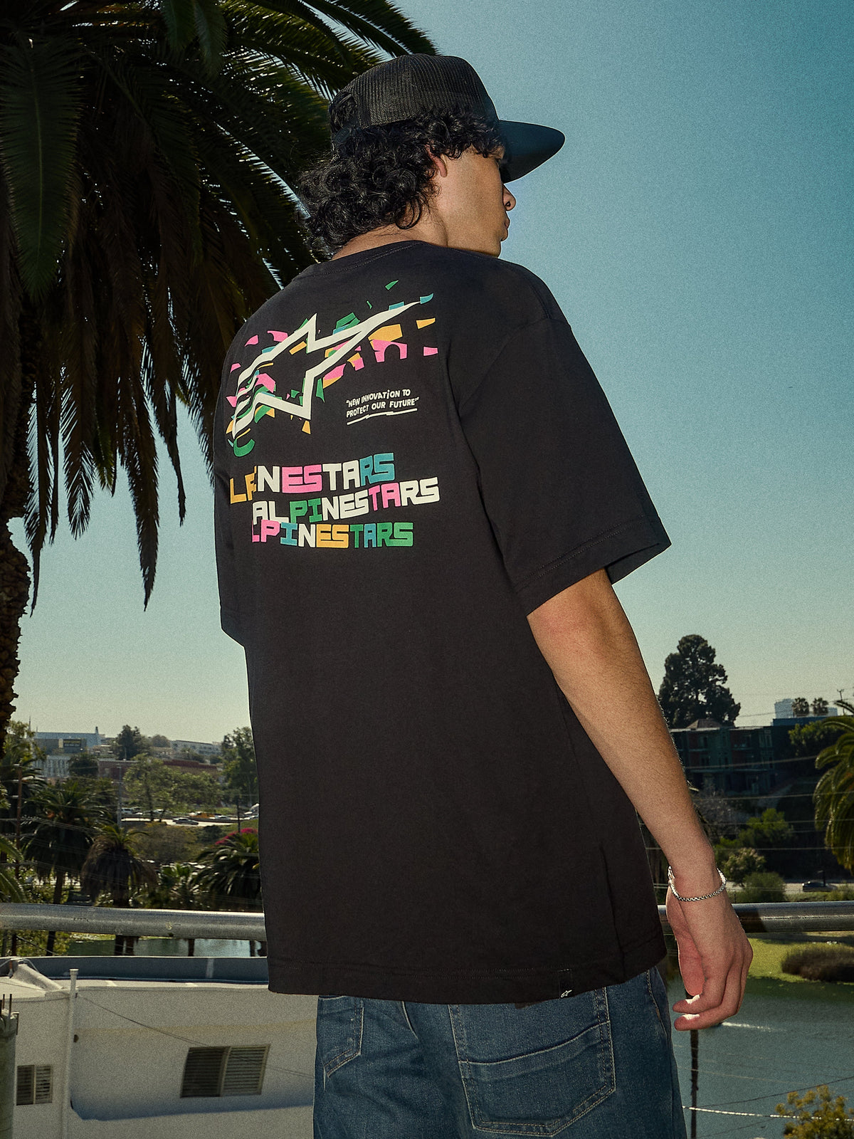 Alpinestars Wild Oversized CSF Tee, Kurzarm-T-Shirt, Schwarz, lebendiges mehrfarbiges Astars-Logo auf der Rückseite, Oversized-Passform, lässige Streetwear-Lifestyle-Aufnahme mit Palmen