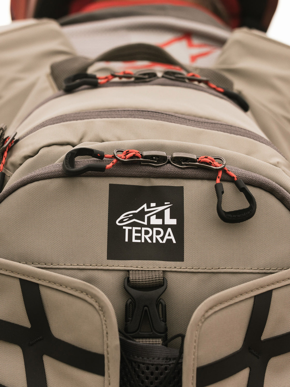 Alpinestars AT-15 Rucksack, Aluminum Walnut, Nahaufnahme des All Terra Logo-Patches auf beigem technischem Textil, mit orangefarbenen Kordel-Reißverschluss-Zügen und robusten Konstruktionsdetails