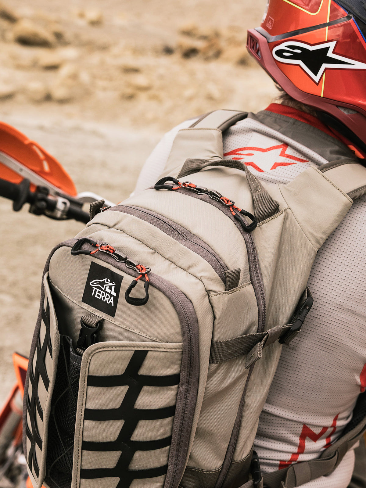 Alpinestars AT-15 Rucksack, Adventure-Motorradrucksack, Aluminum Walnut und Schwarz, getragen von einem Fahrer in einer Wüstenlandschaft, mit roten Reißverschluss-Zippern, TERRA-Logo-Patch und schwarzem Gurtband-System, kombiniert mit passendem roten Helm und weißem Alpinestars-Trikot