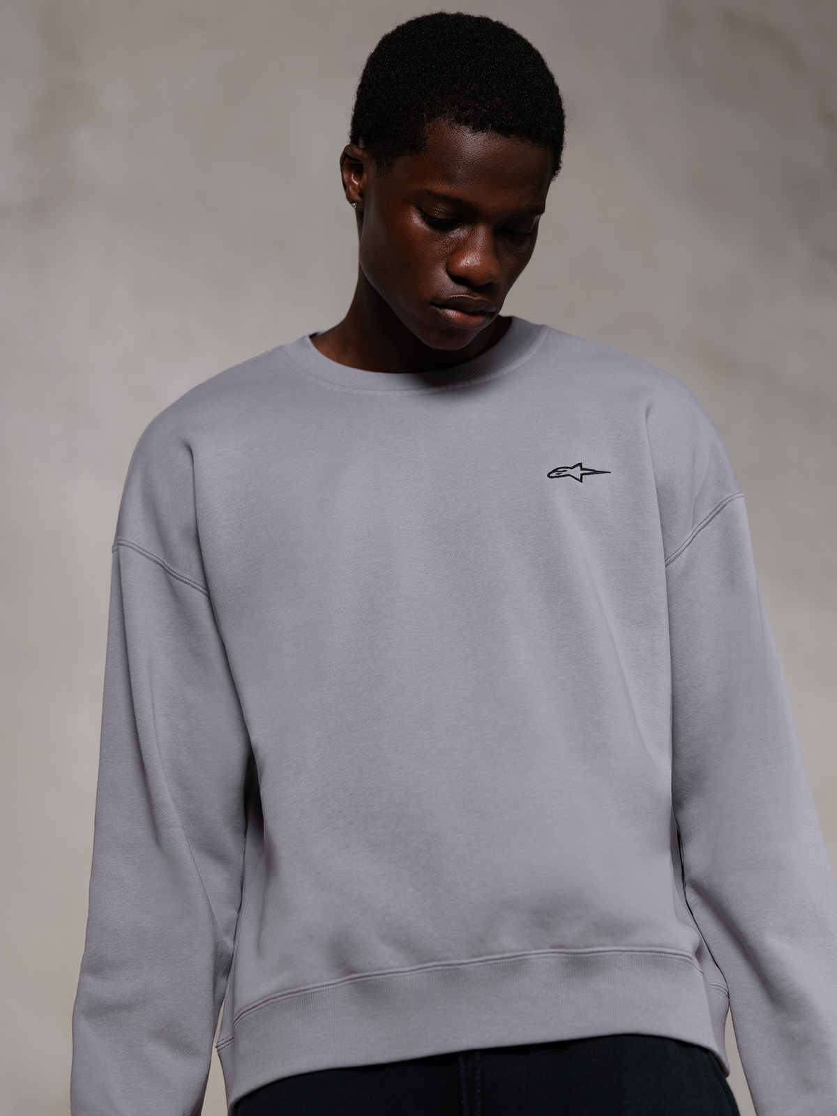 Alpinestars Essentials Ageless Crew, hochwertiges Rundhals-Sweatshirt in Silber, mit einem kleinen schwarzen Alpinestars-Logo auf der Brust, von einem Model getragen, lässige Lifestyle-Streetwear mit weicher Fleece-Textur.