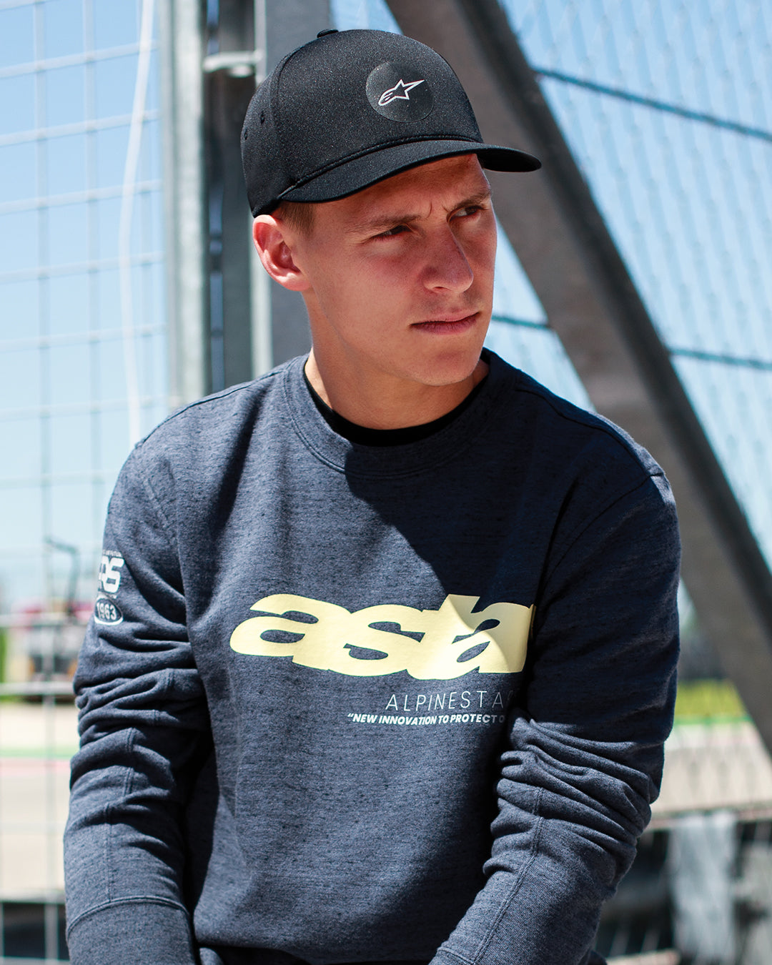 Fabio Quartararo trägt die Alpinestars Apex Mütze in Schwarz, Cap mit strukturierter Krone, gebogenem Schirm und kreisförmigem Stern-Logo, kombiniert mit einem dunkelgrau melierten Sweatshirt mit gelbem Branding, lässiger Motorsport-Lifestyle