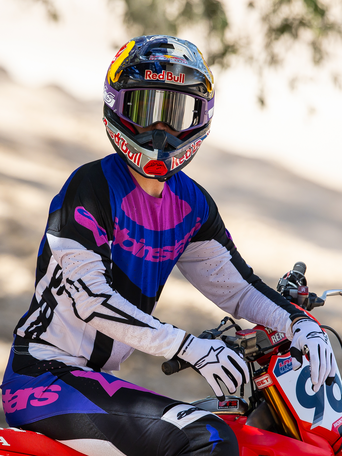Motocross rider on a red dirt bike, Limited Edition HUNTA96 XXV Supertech Vision Goggles in purple and white with silver mirror lenses, matching purple black and white Alpinestars jersey and pants, Red Bull helmet, white gloves, coordinated professional motocross racing gear.