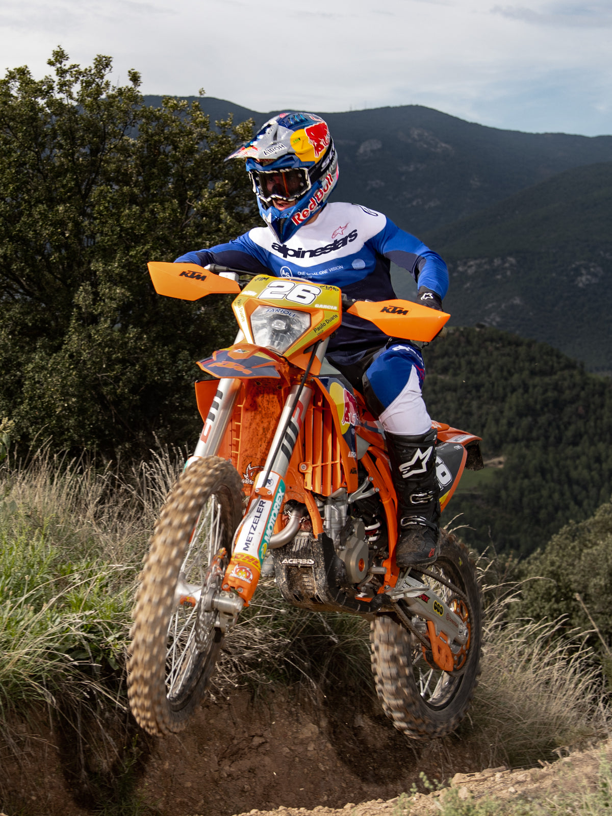 Motocross-Fahrer in Aktion auf einem orangefarbenen KTM-Dirtbike, trägt Alpinestars Pro-Dura Ausrüstung in Navy, Bright Blue und Weiß. Professionelles Offroad-Outfit mit blau-weißem Jersey und passender Hose, Red Bull Helm und schwarzen Stiefeln auf einem Bergpfad.