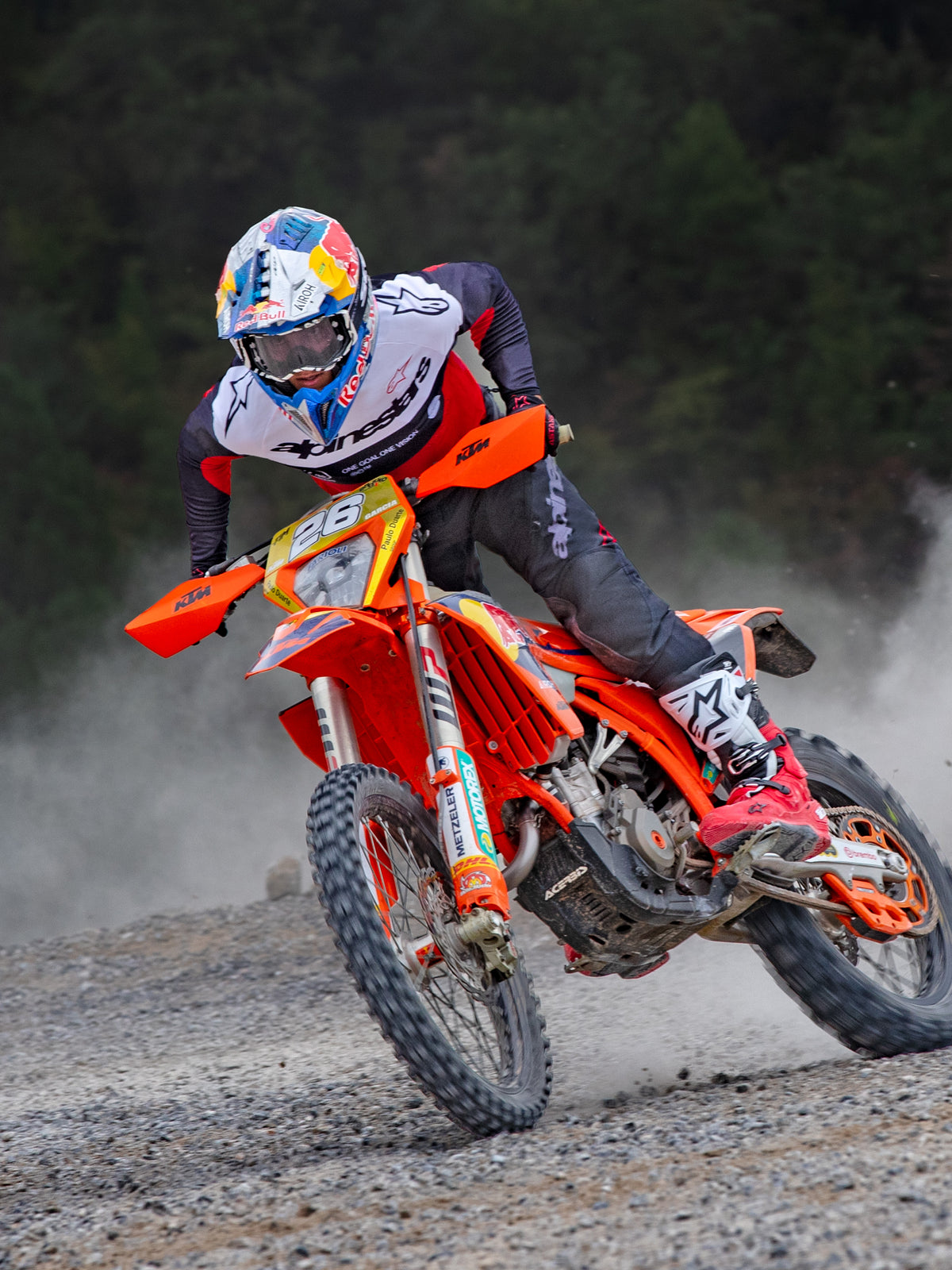 Motocross-Fahrer in Aktion auf einem orangefarbenen KTM-Bike, trägt Alpinestars Pro-Dura Ausrüstung bestehend aus einem rot-weiß-schwarzen Trikot, einer dunkelgrauen Hose, rot-weißen Stiefeln und einem Red Bull Rennhelm, während einer scharfen Kurve auf einem Schotterweg