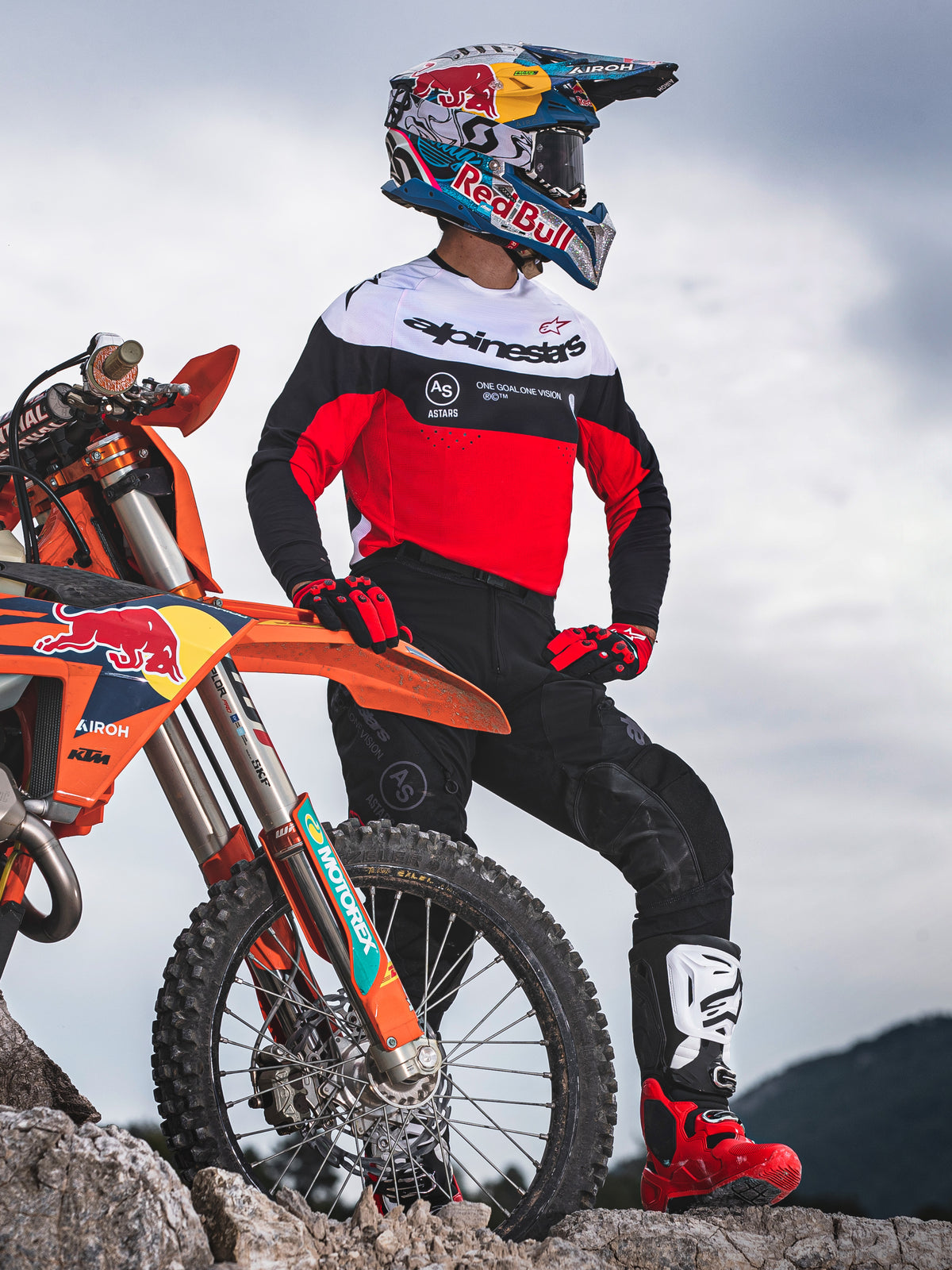 Motocross-Fahrer steht neben einem orangefarbenen Offroad-Motorrad auf felsigem Gelände, trägt Alpinestars Pro-Dura-Ausrüstung mit einem rot, weiß und schwarzen Jersey und einer schwarzen Hose, kombiniert mit rot-schwarzen Handschuhen, weiß-roten Stiefeln und einem Red Bull Helm, komplettes professionelles Enduro-Outfit.