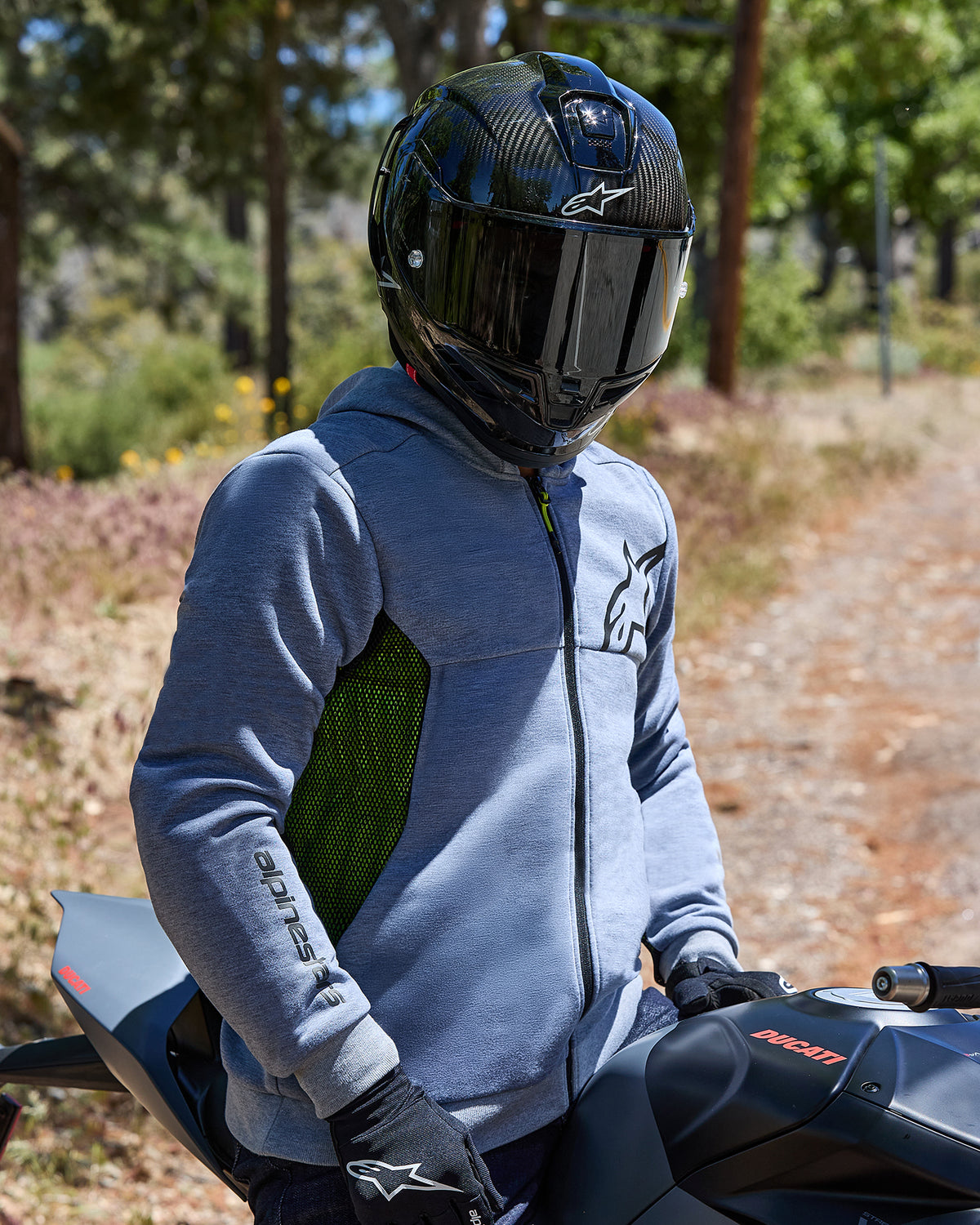 Motorradfahrer im Alpinestars Chrome Air Sport Hoodie in Gray Heather, mit limettengrünen Mesh-Seiteneinsätzen und schwarzen Logos, kombiniert mit einem schwarzen Carbon-Helm und schwarzen Handschuhen, sitzend auf einem Ducati-Motorrad im Freien
