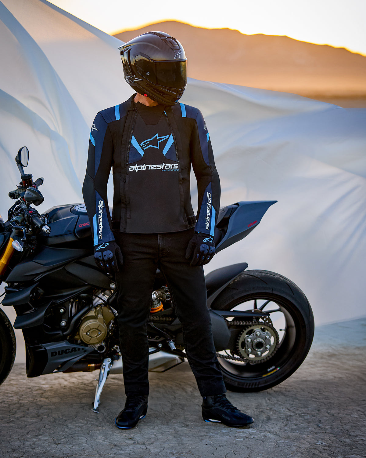 Alpinestars T-Stunt Air Jacke, sportlich geschnittene Textiljacke in Schwarz, Dunkelblau und Hellblau, getragen von einem Fahrer neben einem Ducati-Motorrad, inklusive Carbon-Helm und schwarzer Fahrausrüstung, vor einer Wüstenlandschaft bei Sonnenuntergang.