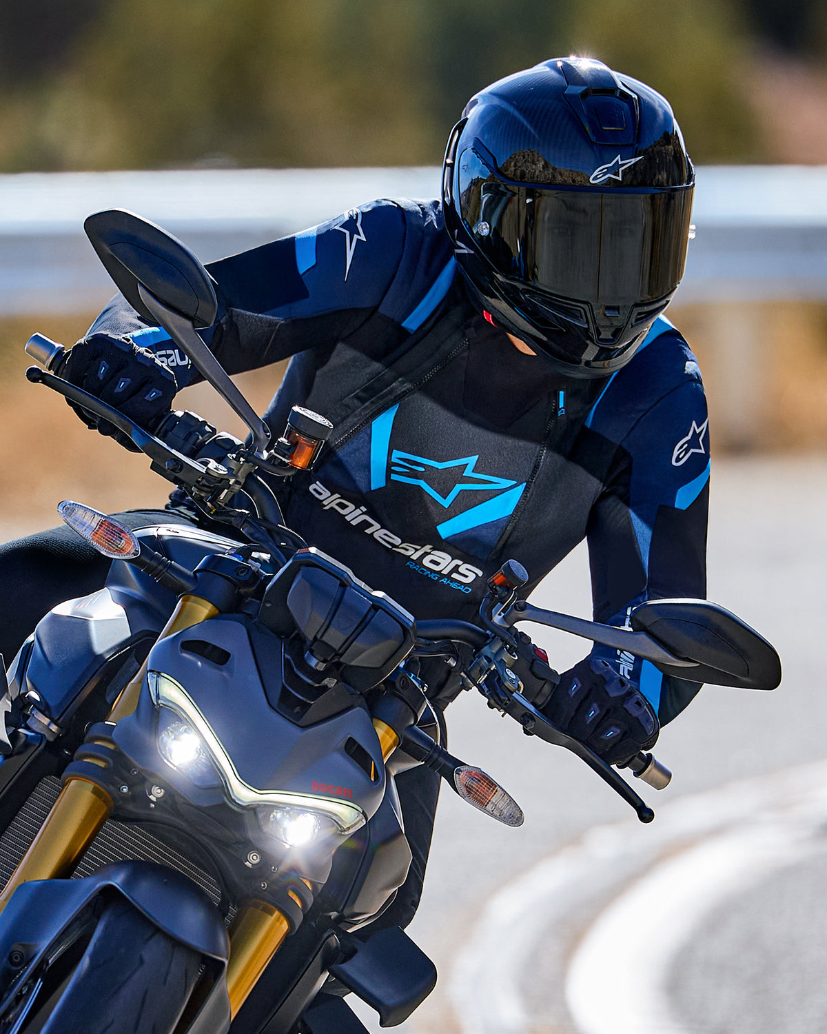 Motorradfahrer in Kurvenlage mit der Alpinestars T-Stunt Air Jacke in Schwarz, Dunkelblau und Hellblau, kombiniert mit schwarzem Helm und Handschuhen, Action-Aufnahme auf einer asphaltierten Straße, Performance-Sport-Fahrerausrüstung