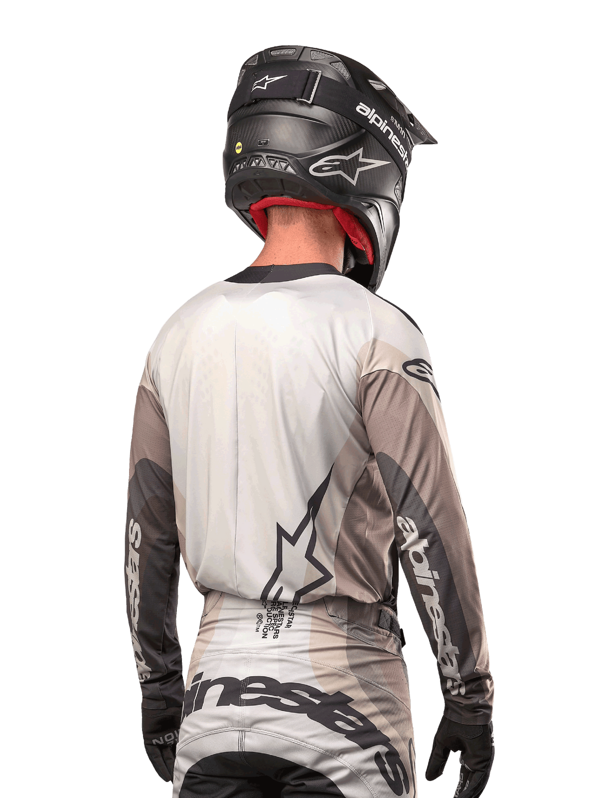 Motocross-Fahrer von hinten im Alpinestars 2024 Techstar Pneuma Jersey in Dark Sand Iron Dust Gray, kombiniert mit einem schwarzen Helm mit Carbon-Finish, mit einem großen Stern-Logo auf dem Rücken und Alpinestars-Branding auf den Ärmeln.