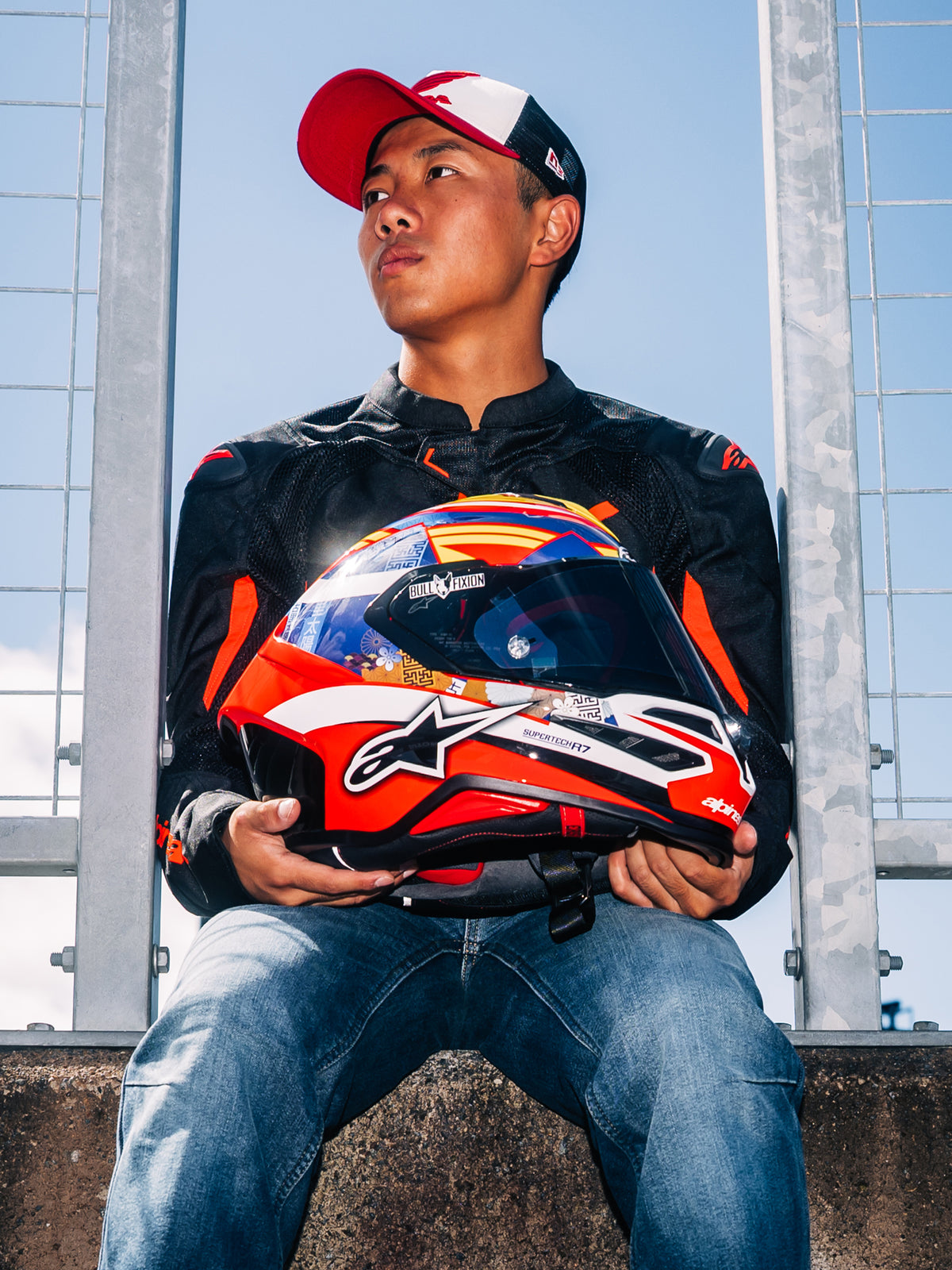 Alpinestars S-R7 Taiyo Furusato Helm, Motorrad-Integralhelm für den Rennsport, rot-weiß-blaue Grafik, gehalten von Fahrer Taiyo Furusato in schwarzer Jacke, Außenaufnahme an der Rennstrecke.