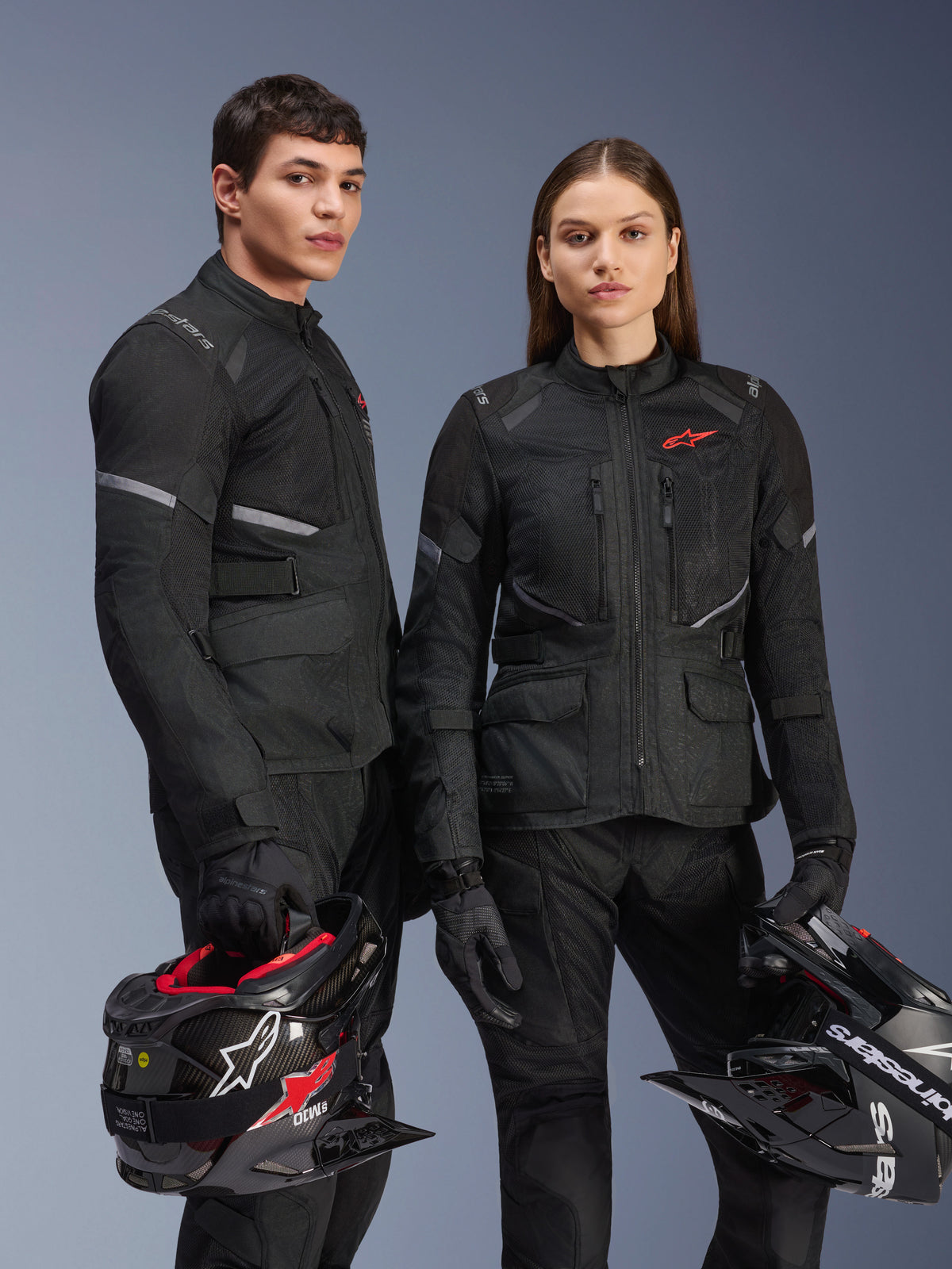 Männliche und weibliche Models tragen Alpinestars Stella Andes Air Drystar® Jacken in Schwarz, Adventure-Motorradtouren-Ausrüstung mit roten Stern-Logos, Mesh-Belüftungseinsätzen und technischer Textilkonstruktion, Models halten Adventure-Helme in einer Studioaufnahme