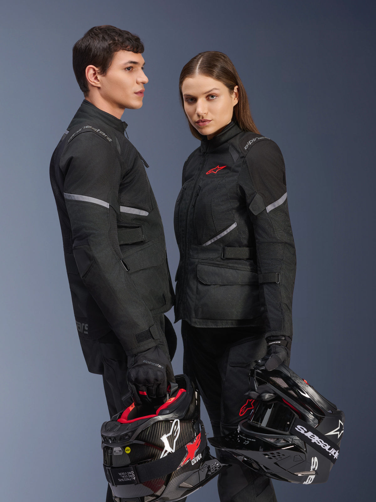 Männliche und weibliche Fahrer in Alpinestars Motorradausrüstung, mit der Stella Andes Air Drystar® Jacke in Schwarz, passender Textilhose und Handschuhen, beide halten schwarz-weiße Helme, Studioaufnahme von koordinierter Adventure-Touring-Bekleidung