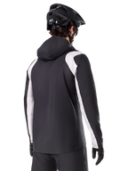 Alpinestars A-Dura Regenjacke, Mountainbike-Regenjacke, Schwarz mit weißen Ärmeleinsätzen, Rückansicht eines Fahrers mit schwarzem MTB-Helm und Handschuhen, technische wasserdichte 2,5-Lagen-Konstruktion, Mountainbike-Bekleidung